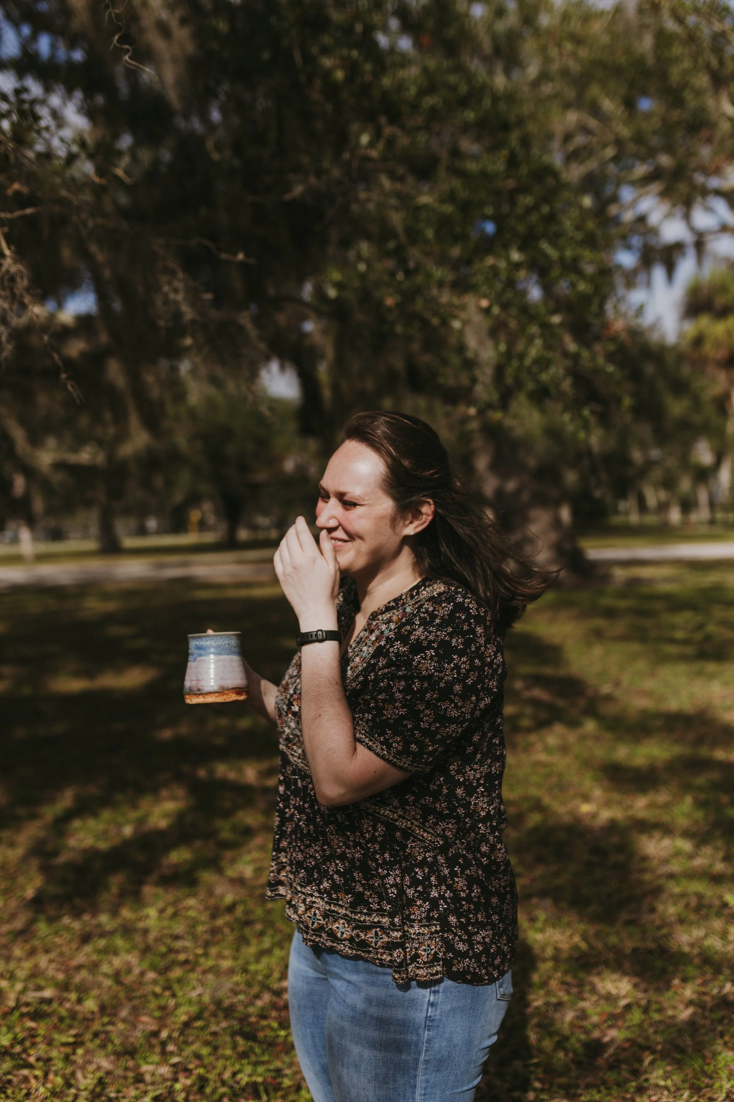 A woman standing outdoors on grass, holding a cup, with her hand near her face, smiling, and her hair blowing in the wind, surrounded by trees.