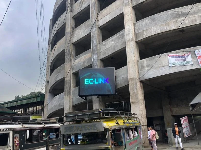 A partially constructed concrete building with open floors and a digital sign that reads 'EC-LINK' mounted on the building facade. A yellow and black jeepney vehicle is parked in front, with a few people nearby.