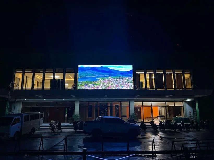 A modern two-story building with large glass windows, illuminated from inside, featuring a large outdoor digital screen displaying a landscape view of mountains, a lake, and a town at night. Several vehicles are parked in front of the building.