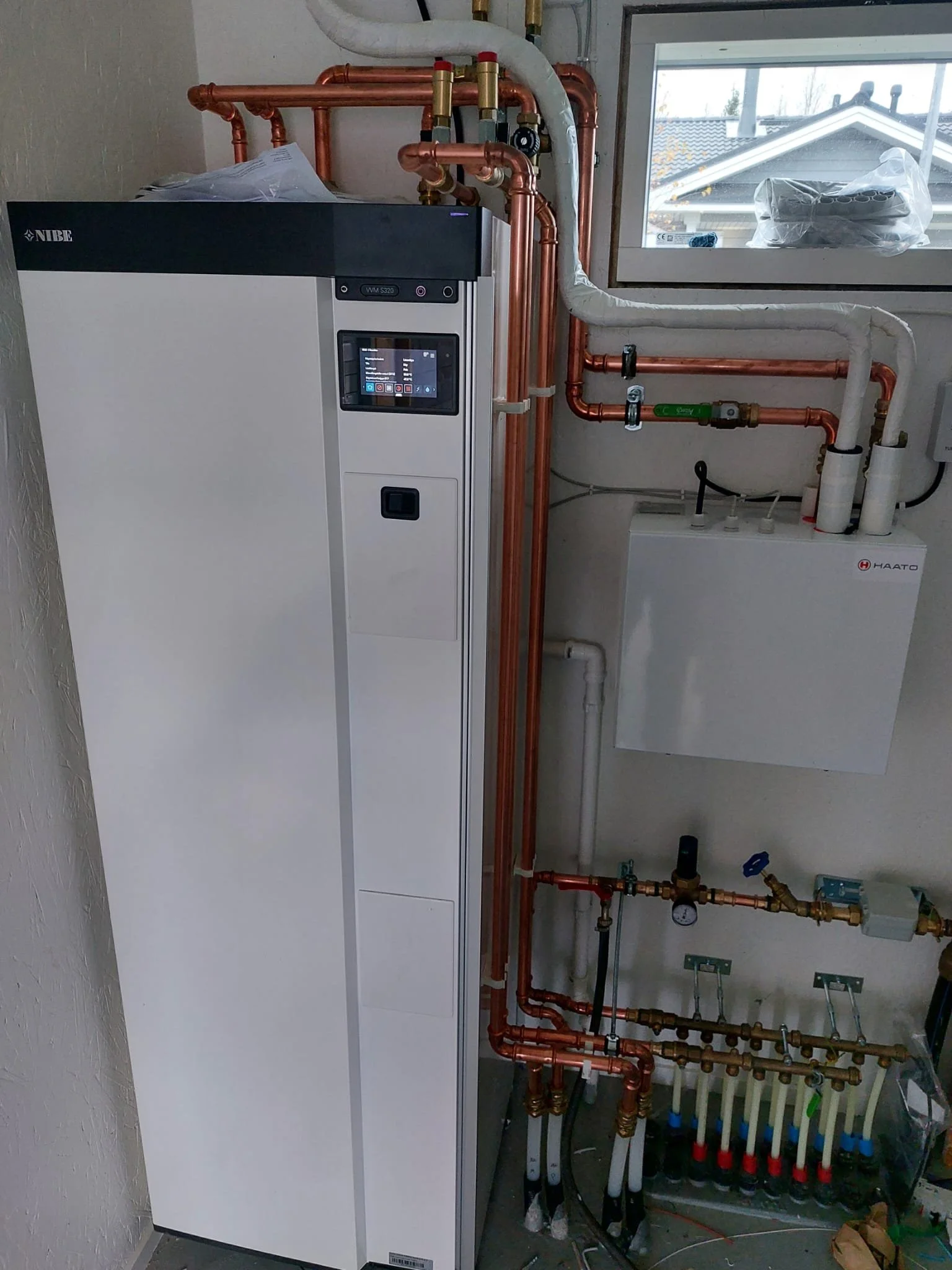 Lämpöpumppu ja putkisto sisätiloissa, seinällä ohjauspaneeli ja ikkuna.