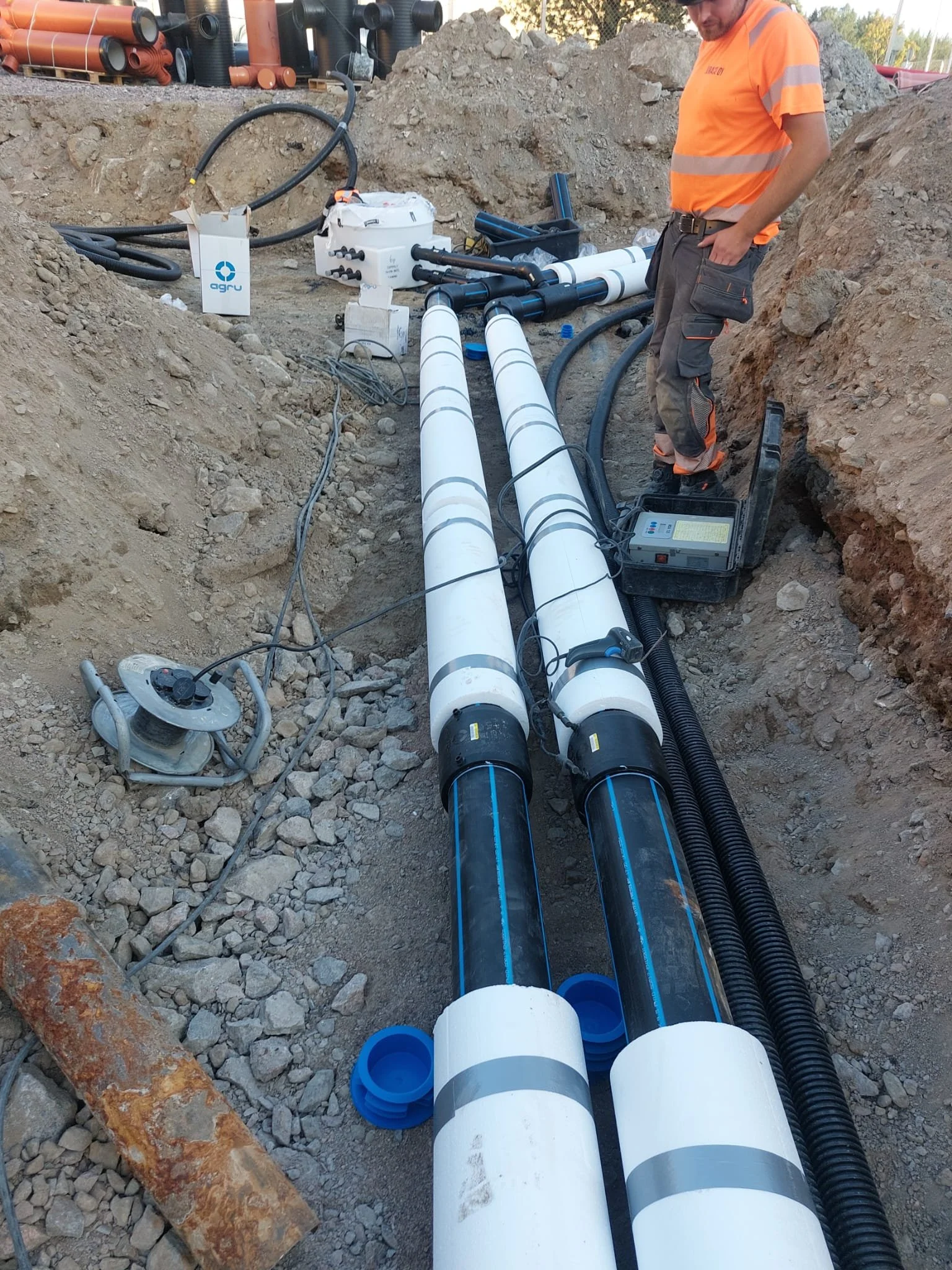 Rakennustyömaa, jossa kaksi suurta putkea kaivannossa ja henkilö työvaatteissa tarkastamassa niitä.