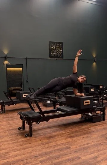 Instructor performing side plank on a Reformer machine in a Pilates studio with dark green walls and wooden flooring.