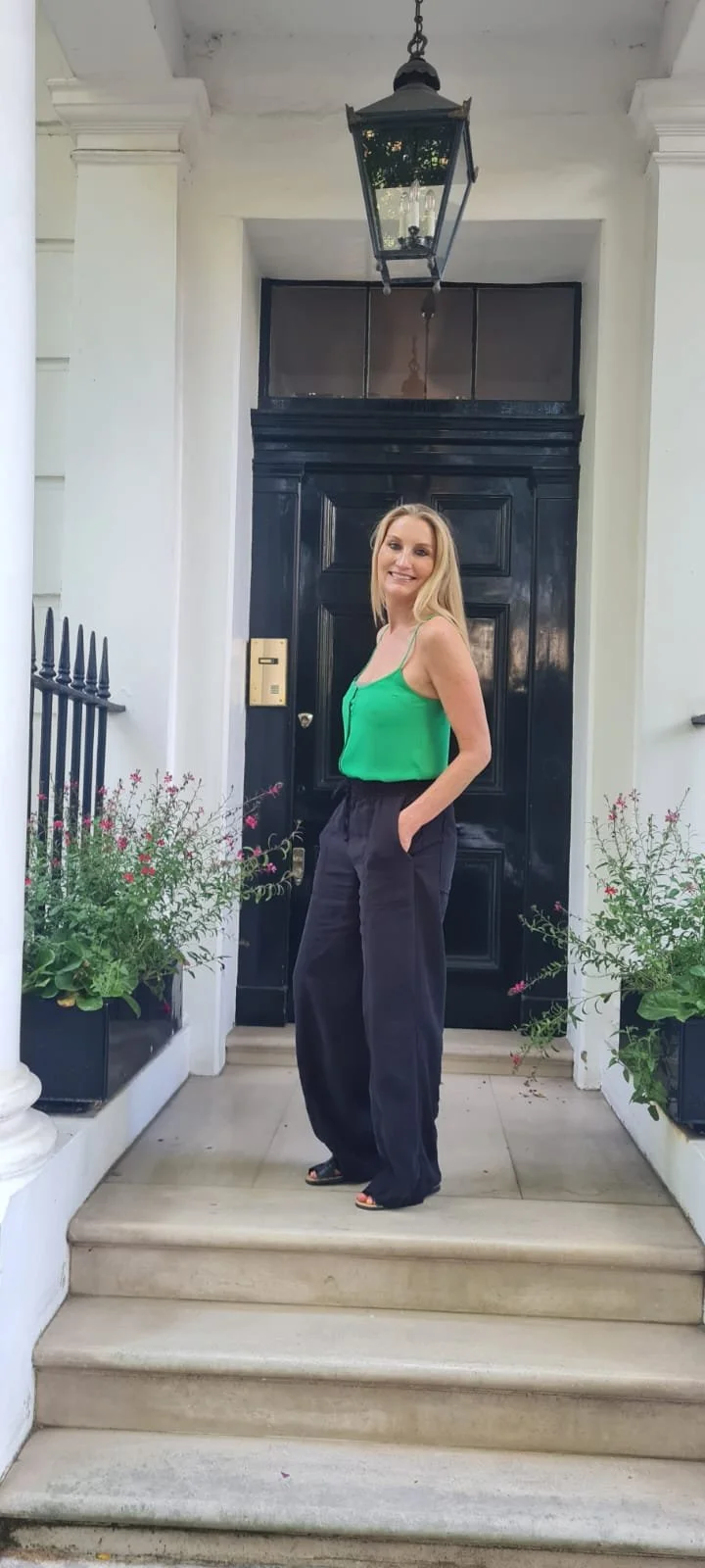 A woman in white workout clothes sitting cross-legged on a yoga mat, stretching with arms raised and smiling.