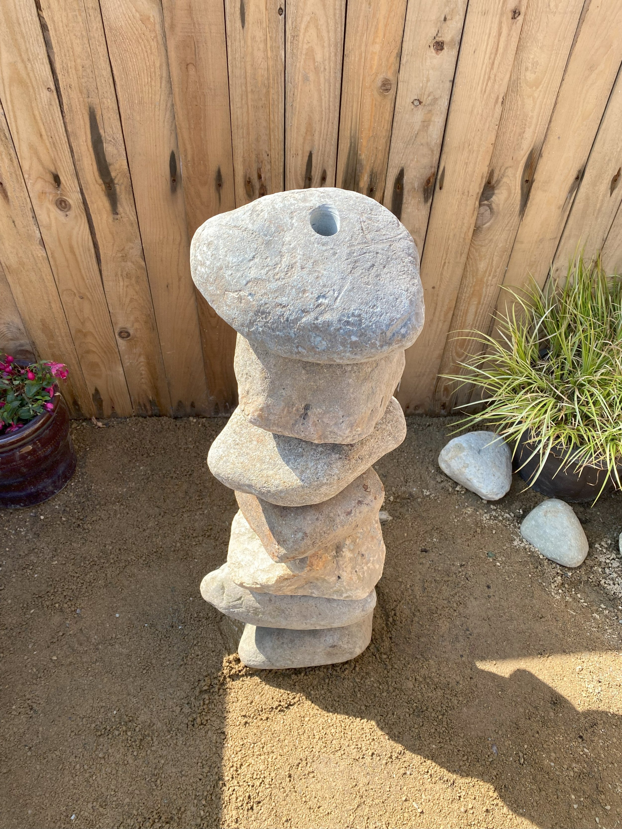 Drilled stone boulders stacked on top of each another in front of fence panelling with plant pots at either side of the stone boulder stack.