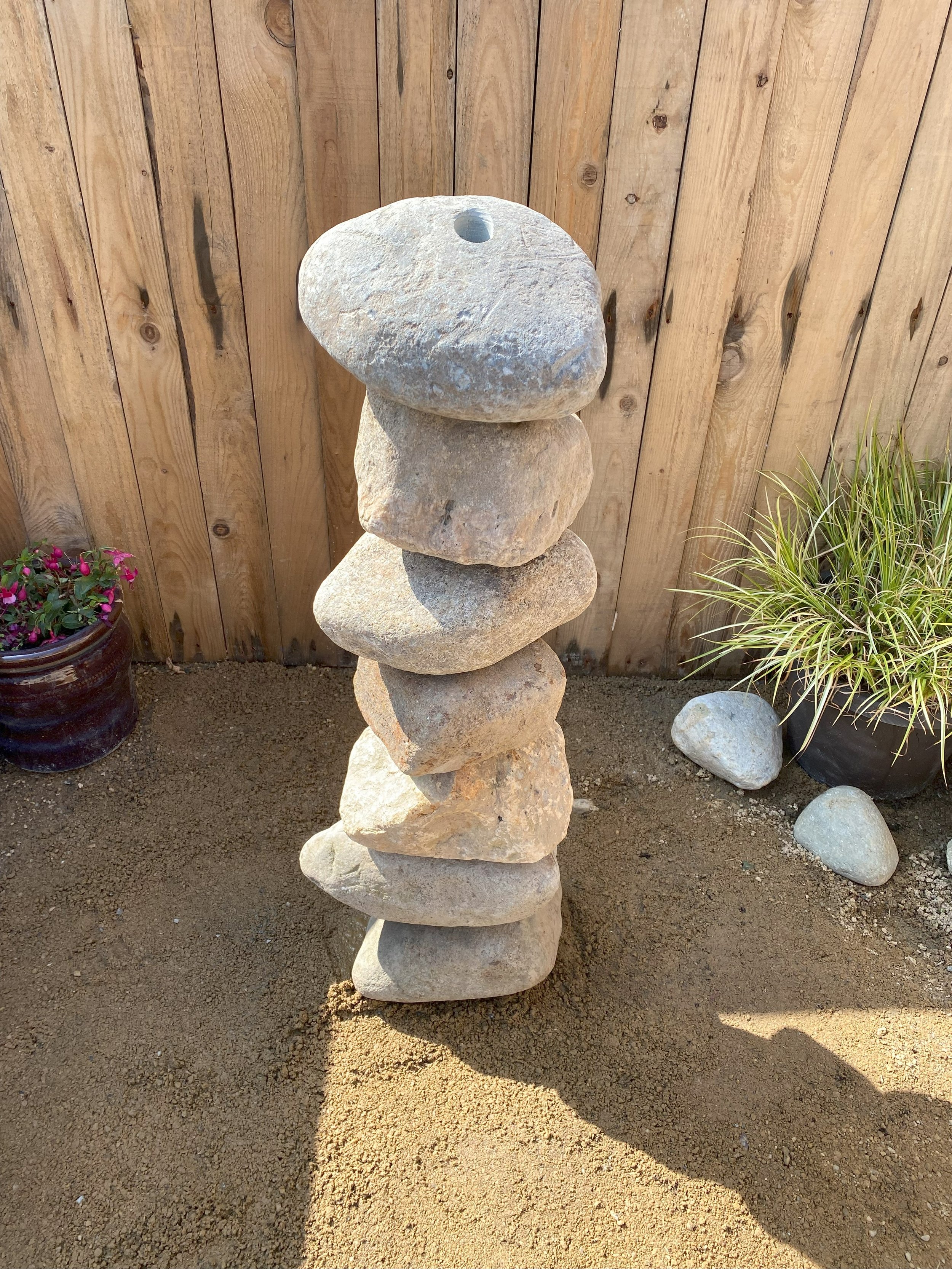 Drilled stone boulders stacked on top of each another in front of fence panelling with plant pots at either side of the stone boulder stack