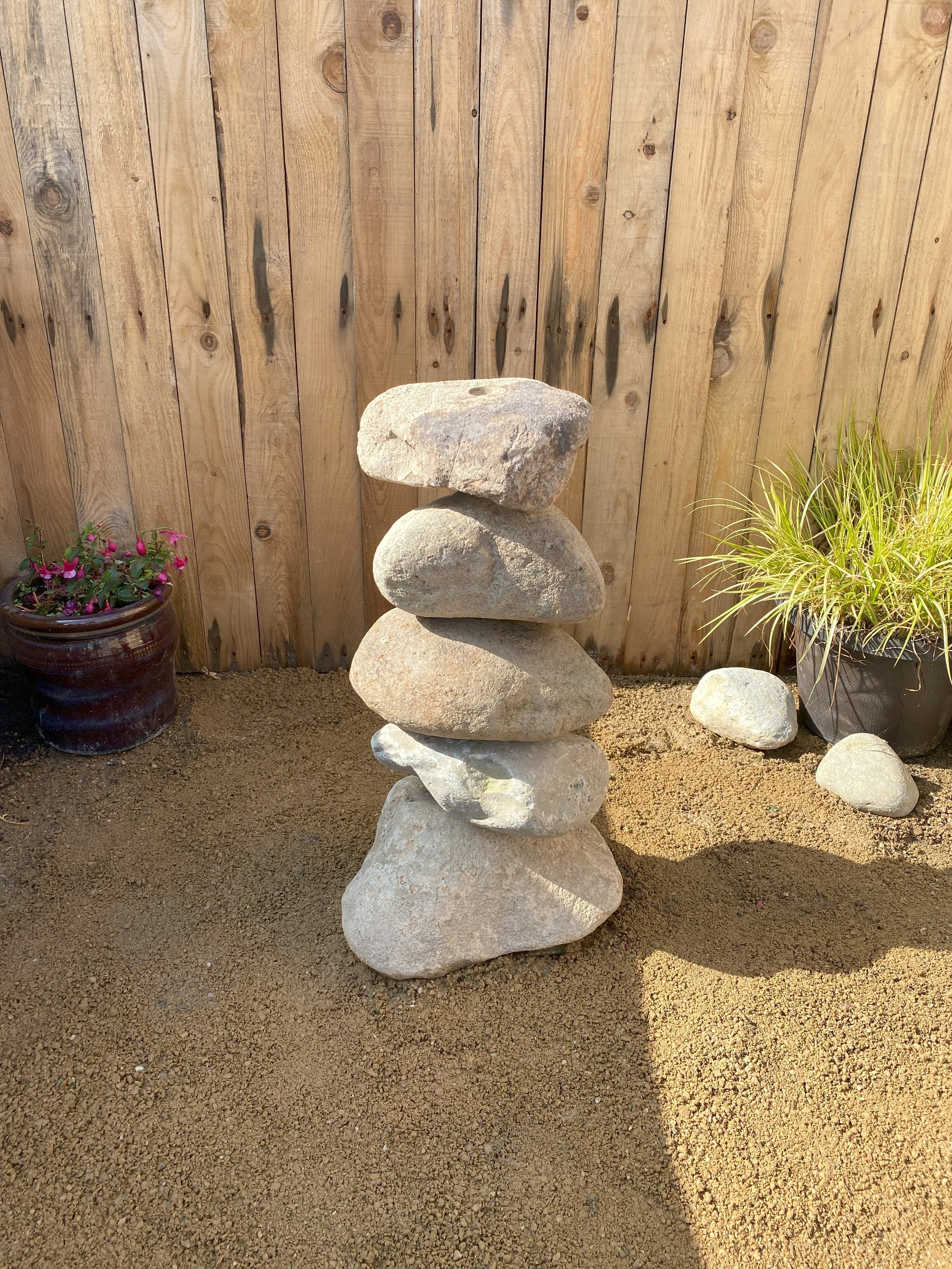 Drilled stone boulders stacked on top of each another in front of fence panelling with plant pots at either side of the stone boulder stack.