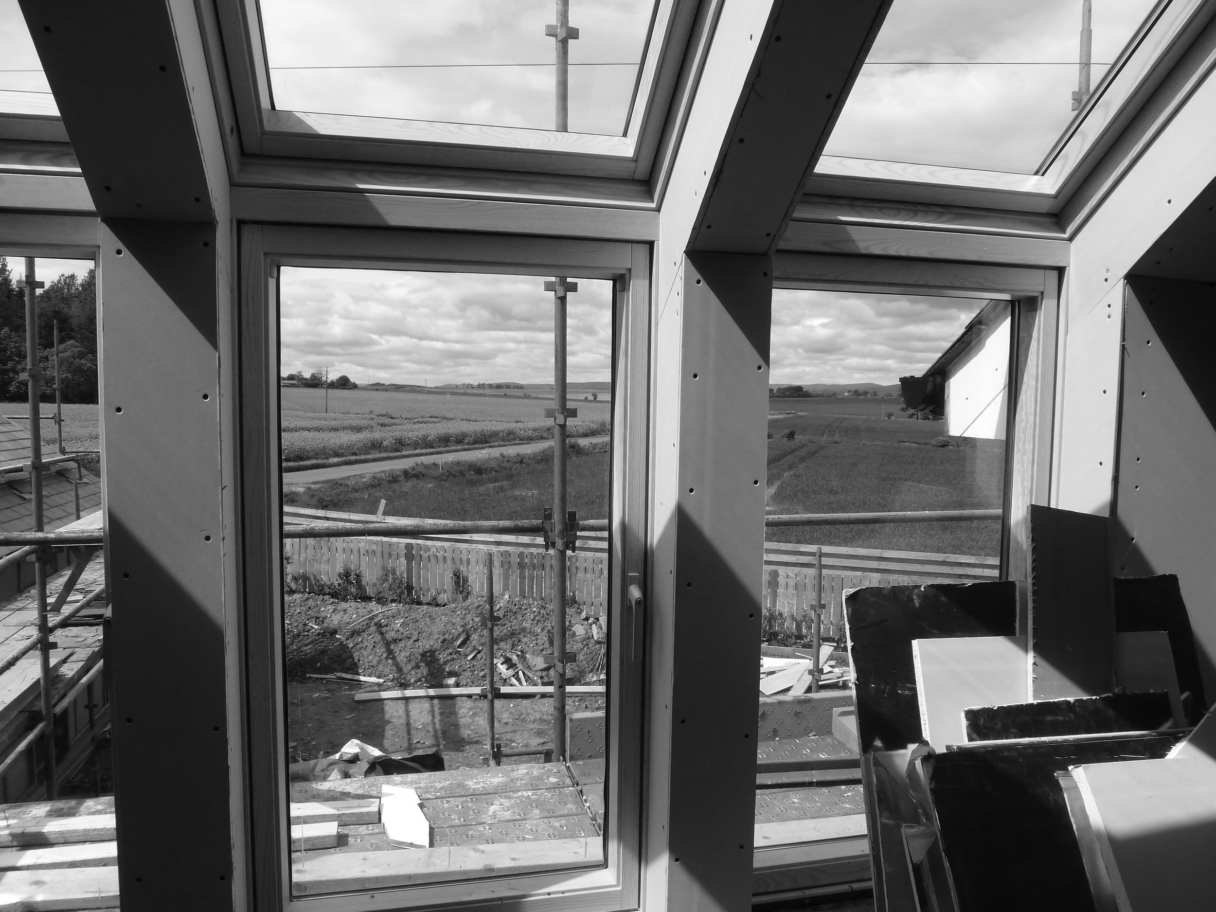 Interior view of a building under construction with large glass windows overlooking a rural landscape with fields and a cloudy sky.