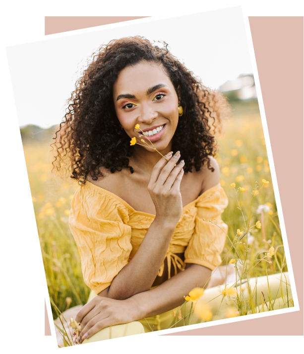 A young woman with curly hair smiling in a yellow off-shoulder dress, sitting in a field of yellow flowers, holding a flower near her face, with a blurred outdoor background.