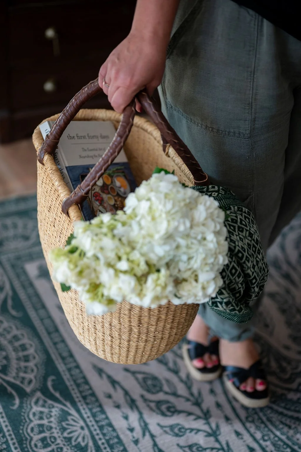 Person holding a wicker basket with white flowers, a book, and magazines inside, standing on a patterned rug.