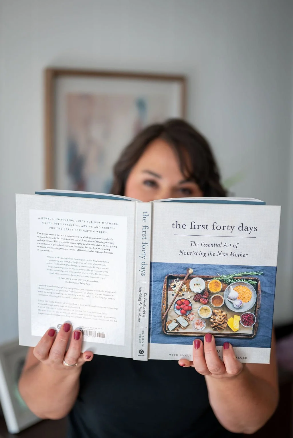 Woman holding a book titled 'The First Forty Days,' obscuring her face, with a blurred framed artwork behind her.