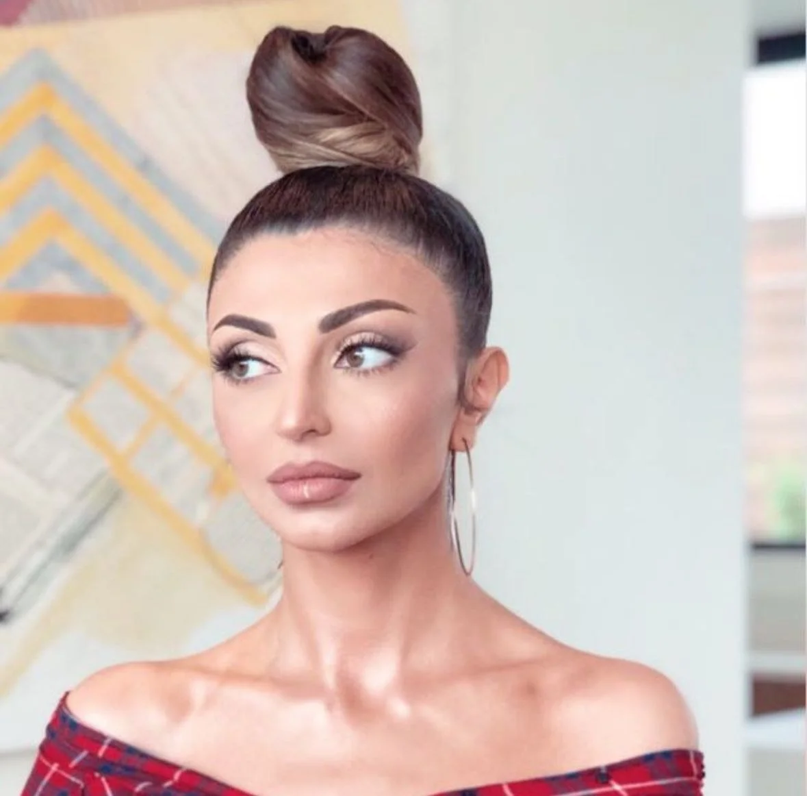 A woman with dark hair styled in a high bun, wearing large hoop earrings and a red plaid off-the-shoulder top. She has makeup with prominent eyebrows, long eyelashes, and nude lipstick, looking to the side with a neutral expression.