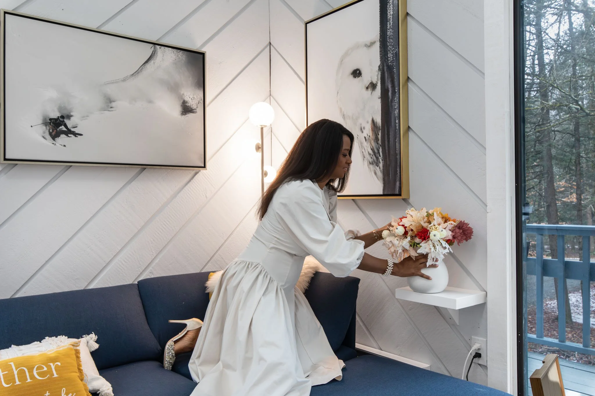 A woman in a white dress arranging a bouquet of flowers in a white vase on a wall shelf inside a modern living room with art and a large window.