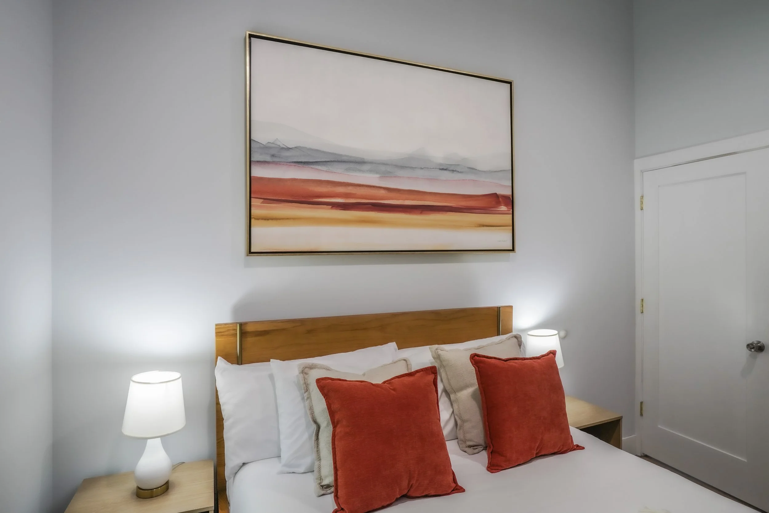 A bedroom with a wooden headboard, white bedding, two white pillows, and two red pillows. There are two lamps, one on each side of the bed, and a large abstract landscape painting hangs above the headboard.
