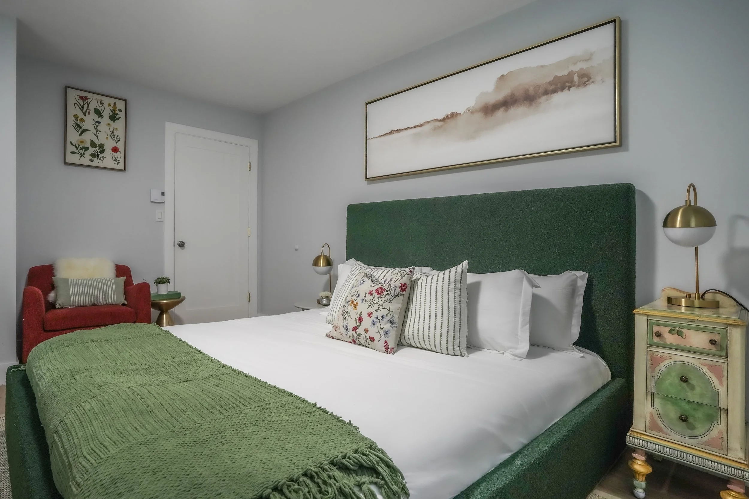 A bedroom with a green upholstered bed, white bedding, decorative pillows, a green blanket, two gold-tone bedside lamps, a colorful painted drawer, a red armchair with a striped pillow and faux fur cushion, and framed artwork on the walls.