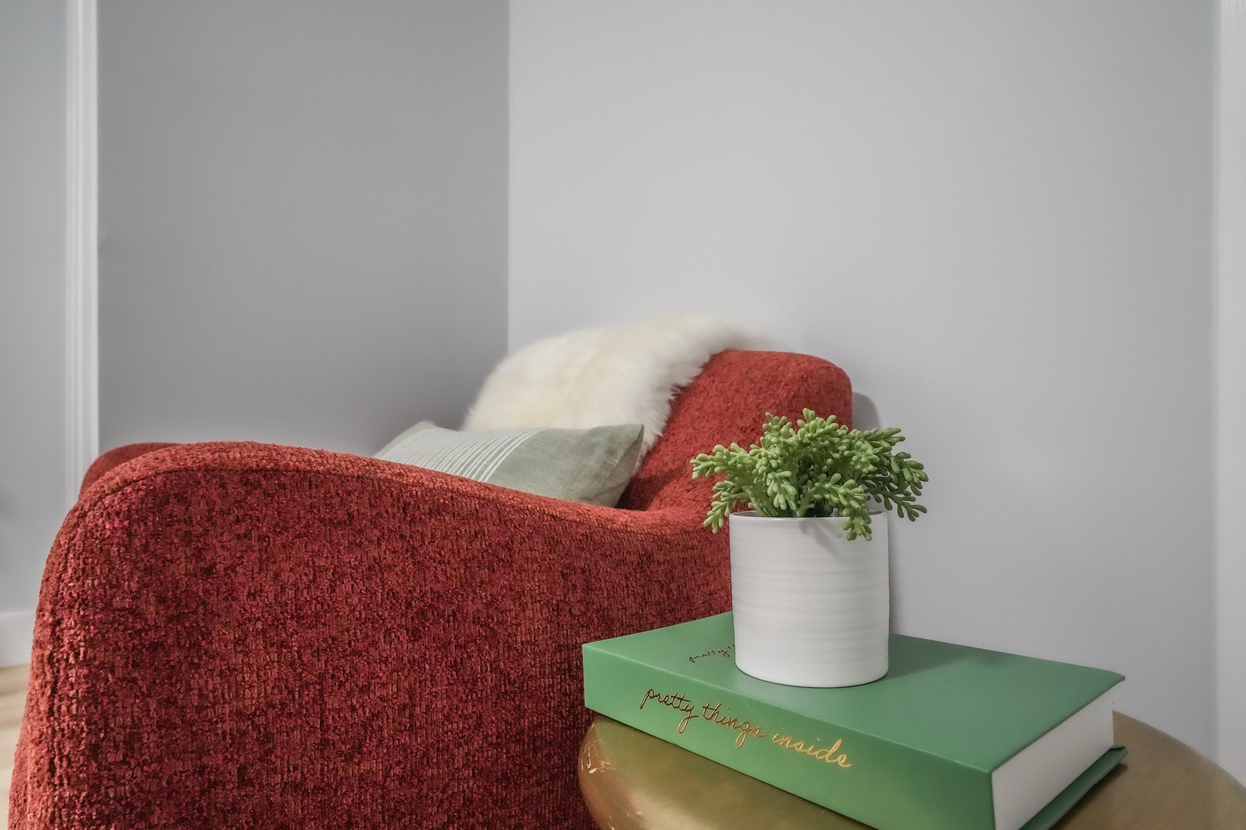 A cozy living room corner with a red upholstered chair, throw pillows, a white furry throw, a green book titled 'pretty things inside' on a wooden side table, and a small green succulent plant in a white pot.