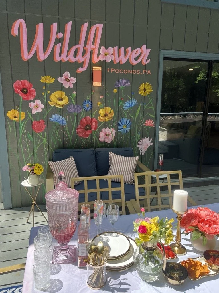 Decorative outdoor patio with floral theme, featuring a wall with a sign that says 'Wildflower Poconos, PA' and painted flowers. There is a table set with drinks, flower arrangements, candles, and plates, and a cozy seating area with a blue couch and striped pillows.