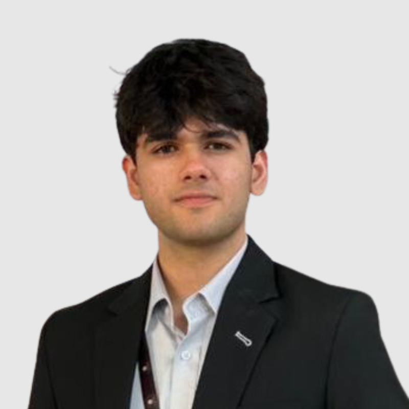A young man with dark hair wearing a black suit and white shirt, standing against a plain light background.