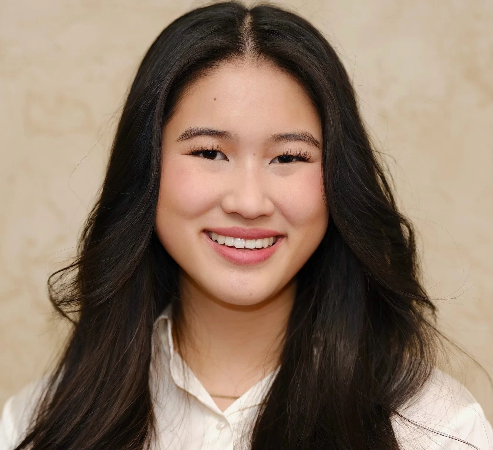Close-up portrait of a young Asian woman with long, wavy black hair, smiling with white teeth, wearing a white shirt, against a beige background.