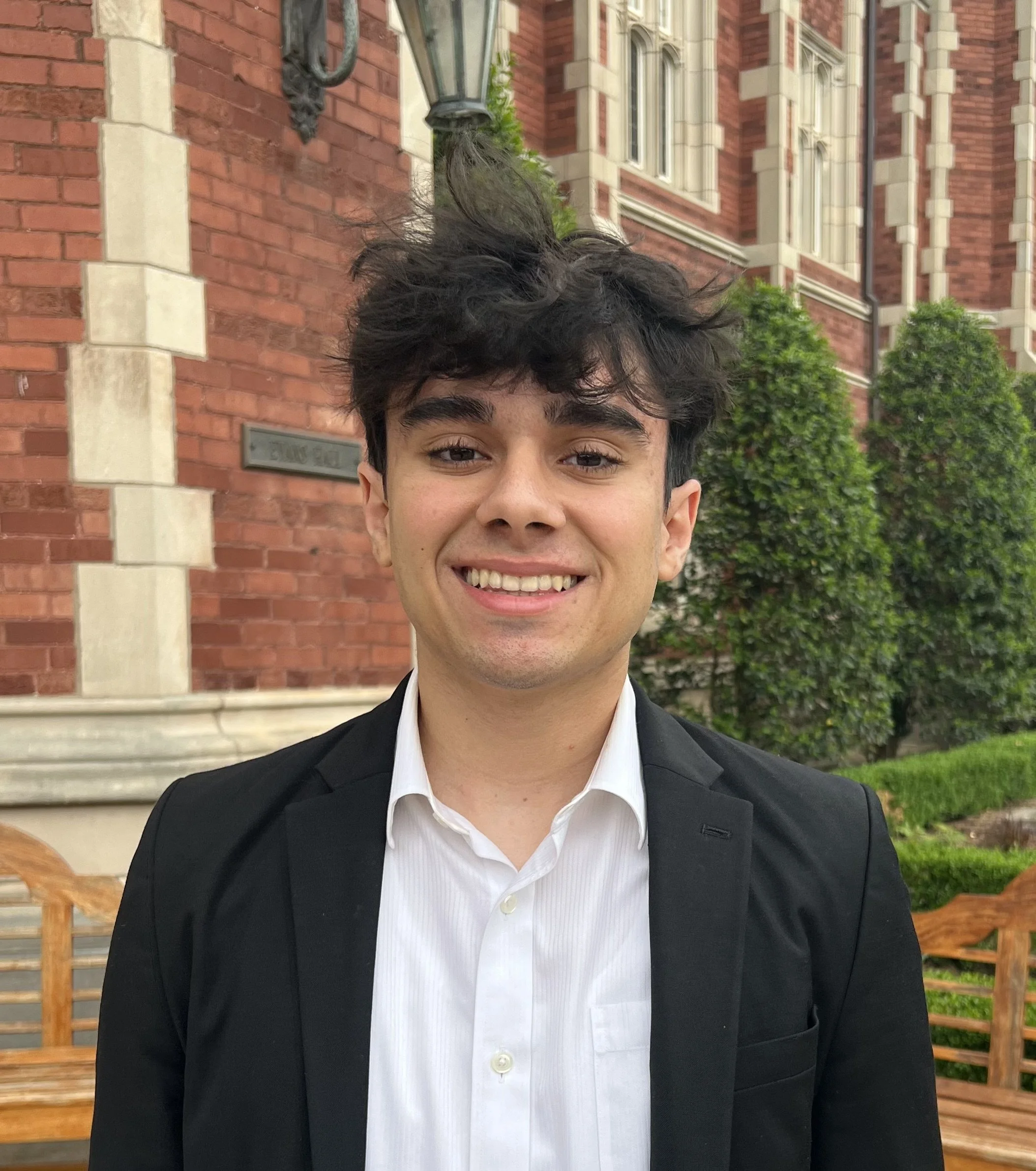 Close-up of a young man with tousled dark hair, wearing a black suit jacket and a white button-down shirt, standing outdoors in front of a brick building with white accents, green bushes, and a wooden bench.