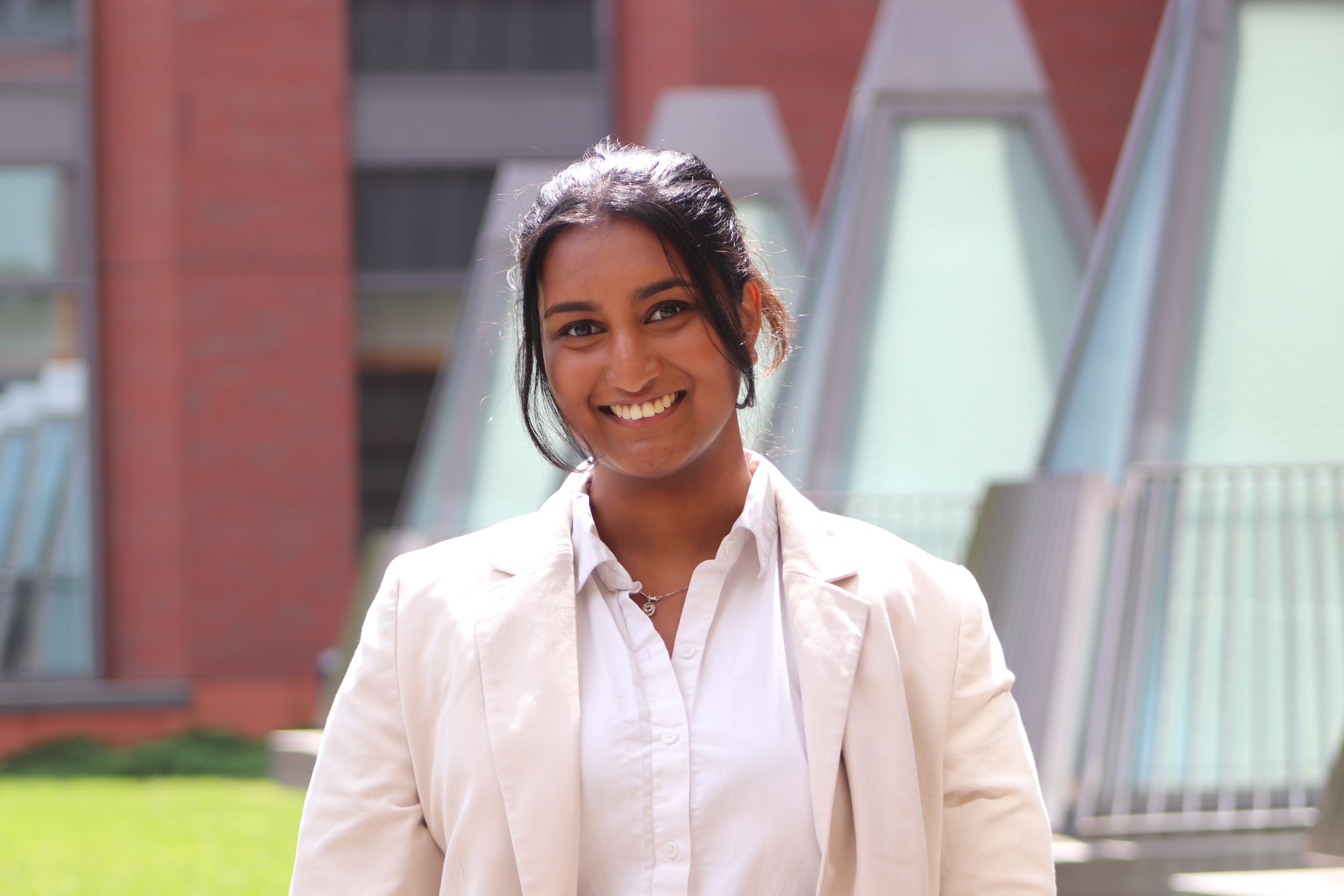 A young woman with dark hair tied back, smiling, wearing a white shirt and beige blazer, standing outdoors near modern buildings with large glass windows.