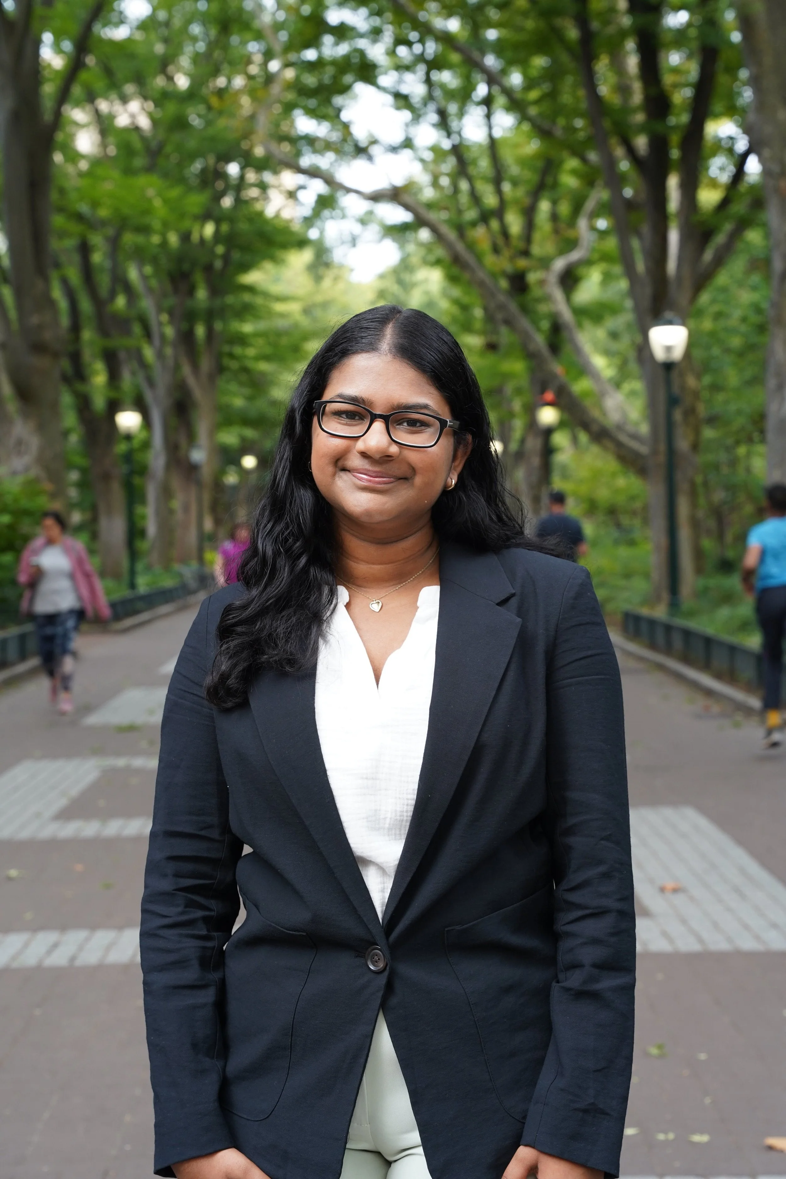A woman with long black hair and glasses, wearing a black blazer and a white blouse, standing outdoors in a park with trees and lamp posts in the background.