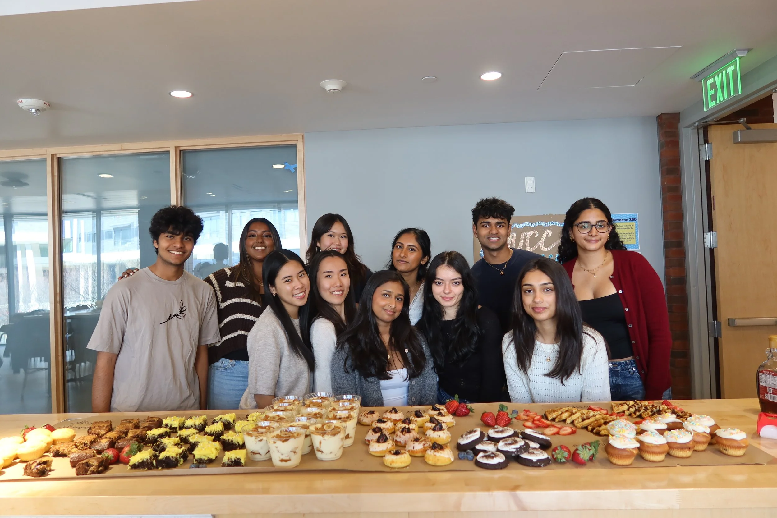 Group of young people smiling behind a table of assorted desserts, including cupcakes, strawberries, and cookies, in a room with glass windows and an exit sign.
