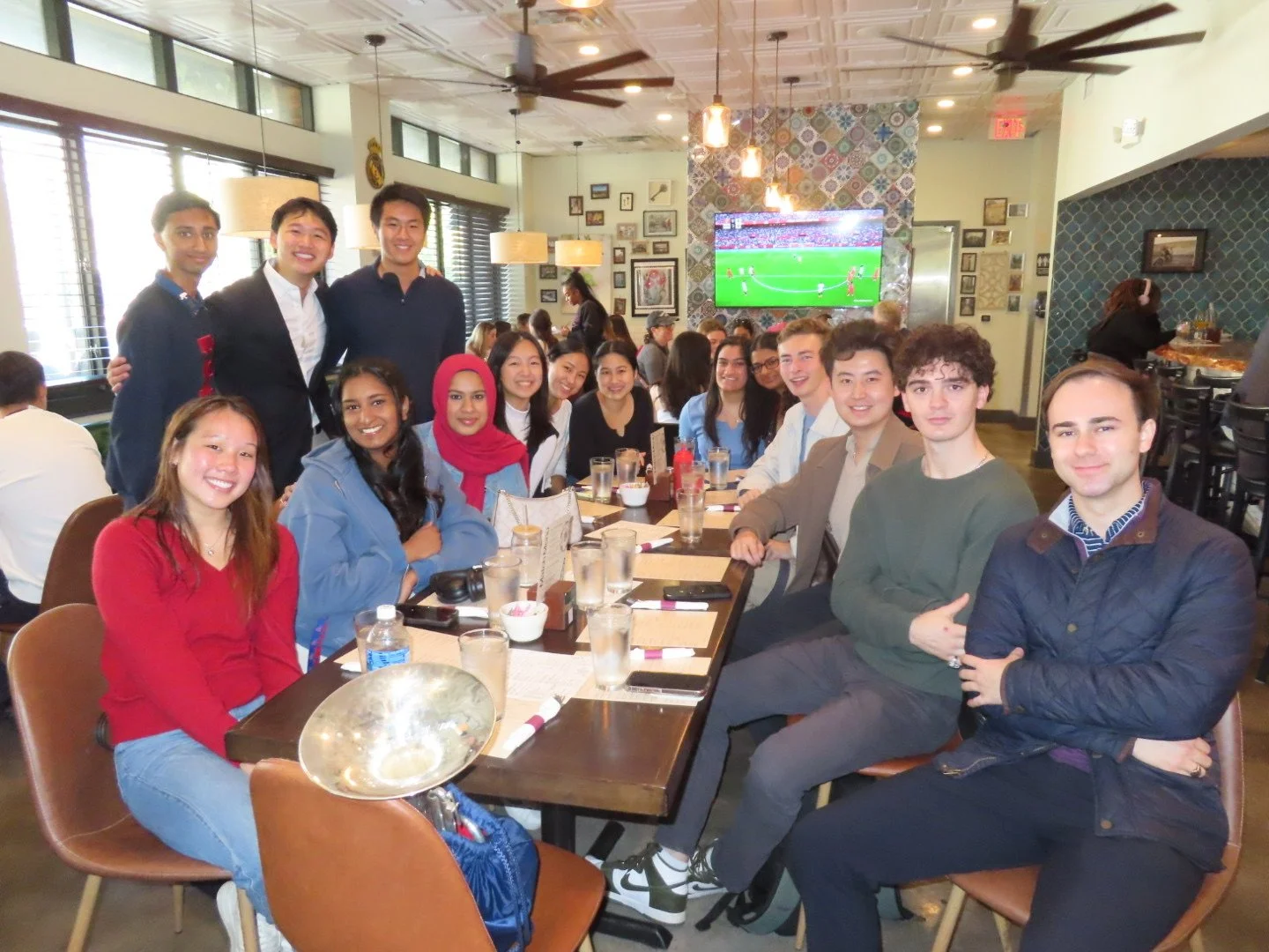 Group of people gathered around a table in a restaurant, watching a soccer game on a television screen.