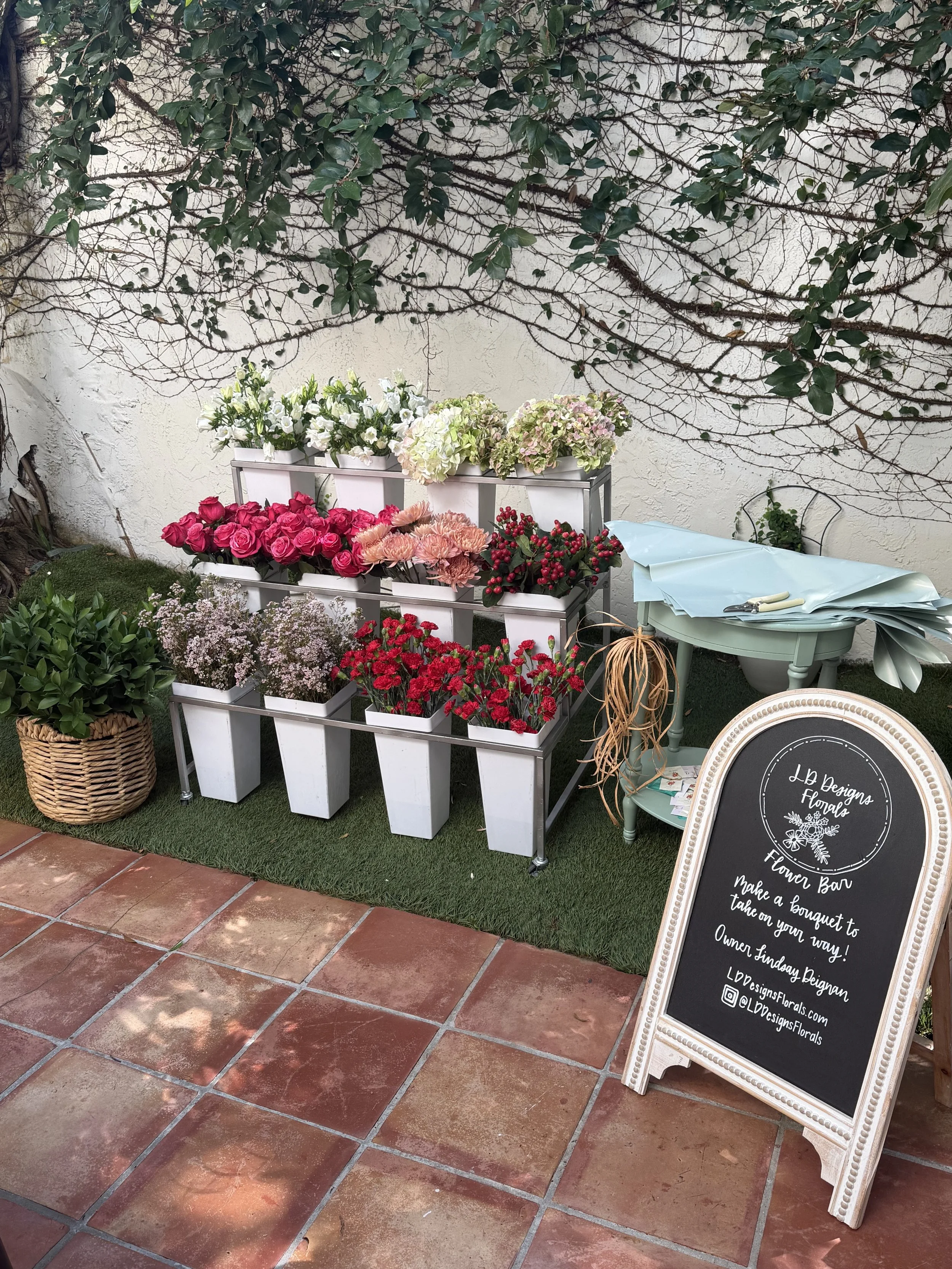 A flower display with various potted flowers including pink roses, white lilies, and hydrangeas arranged on white shelves outdoors. There is a large chalkboard sign with the shop name 'LBD Designs Florals' and information about a flower bar and social media handles, next to a green table with blue paper and tools. The setting has a brick-tiled floor and a white textured wall with climbing greenery.