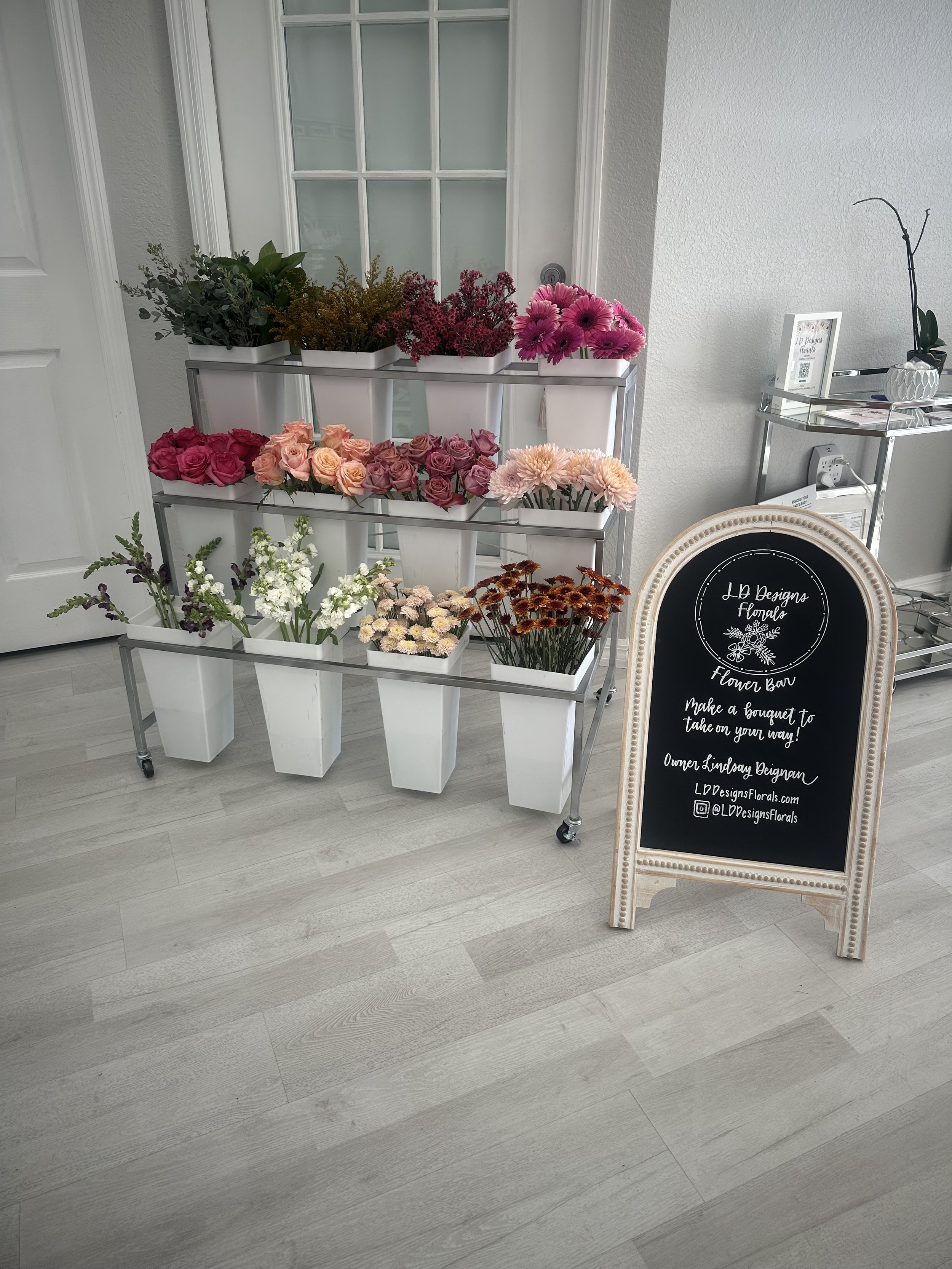 Display of various fresh flowers arranged in white containers on a multi-tiered metal stand, situated next to a black chalkboard sign with white handwritten text promoting a flower bar event.