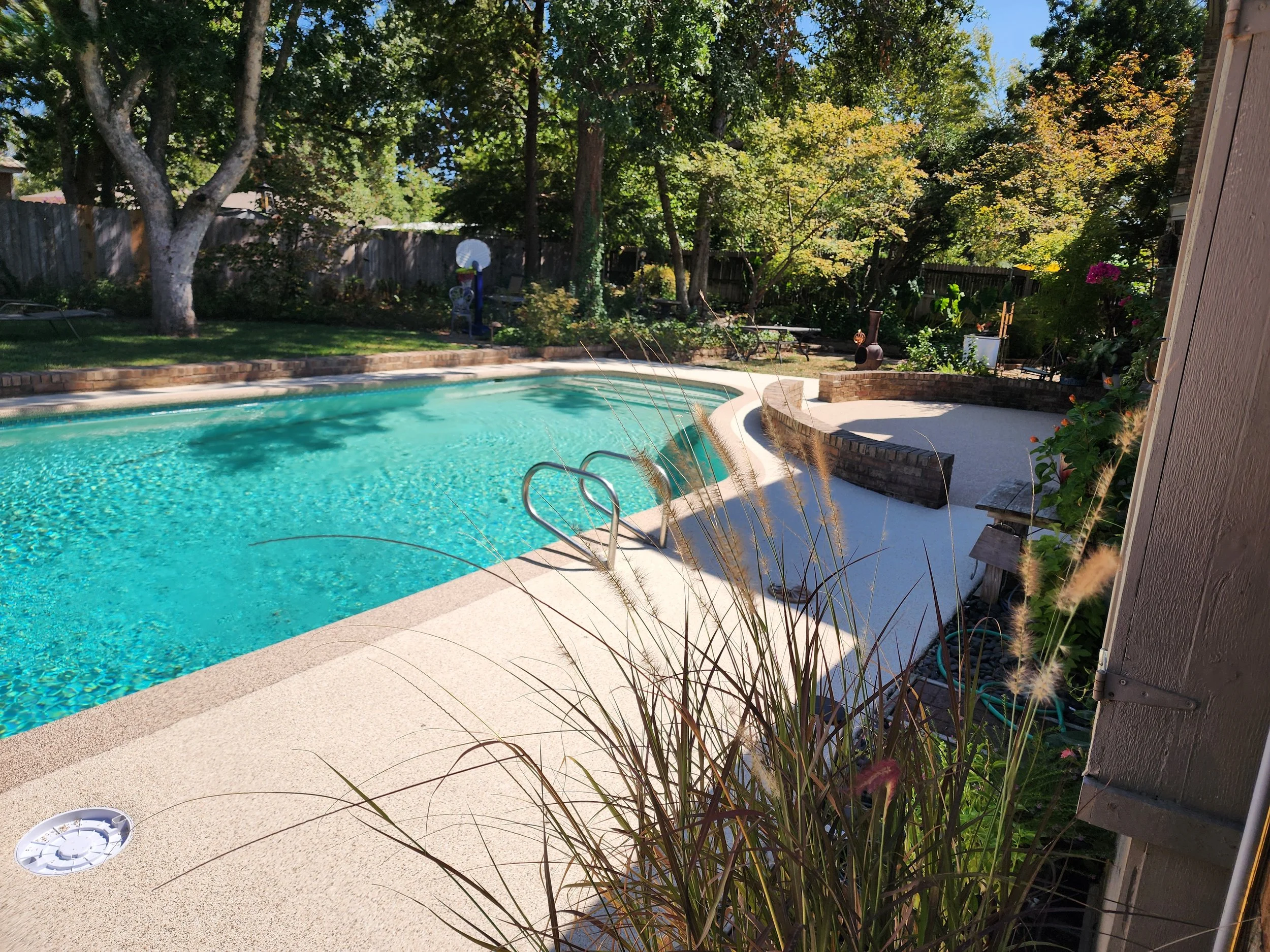 Backyard with a swimming pool, trees, and garden furniture surrounded by a wooden fence.