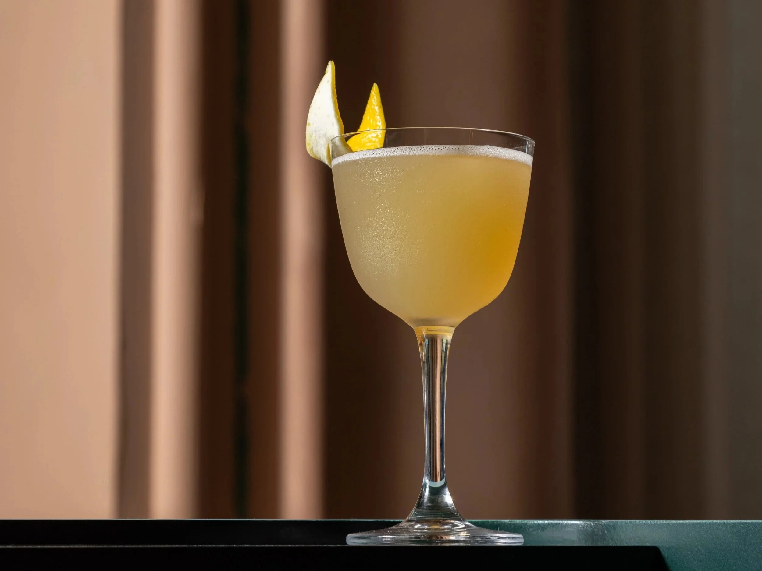 A cocktail in a stemmed glass garnished with lemon and lime wedges against a blurred background