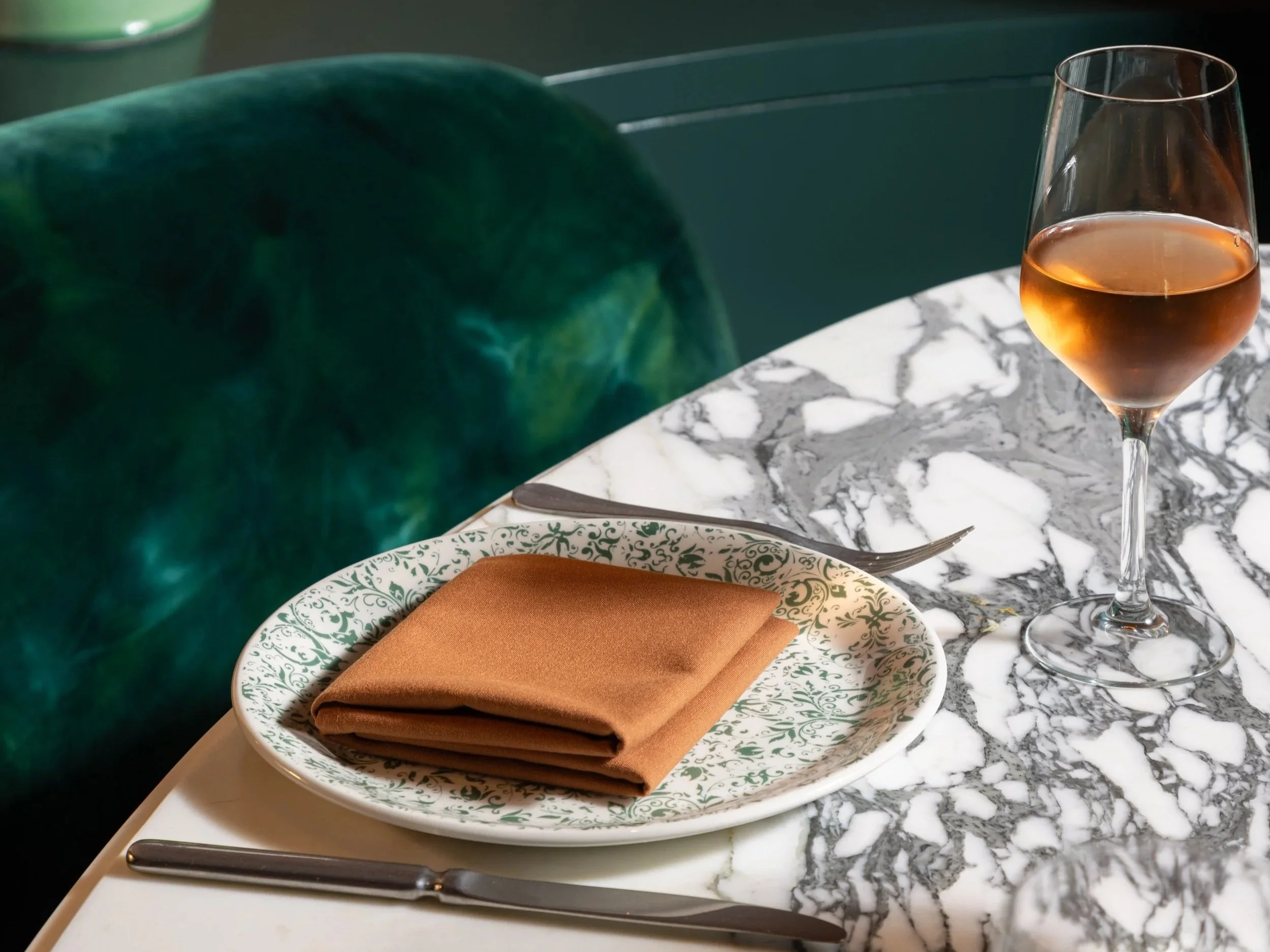 A marble table set with a patterned plate, a brown folded napkin, a knife, a wine glass with rosé wine, and a green velvet chair. There is a green vase and a lamp in the background.