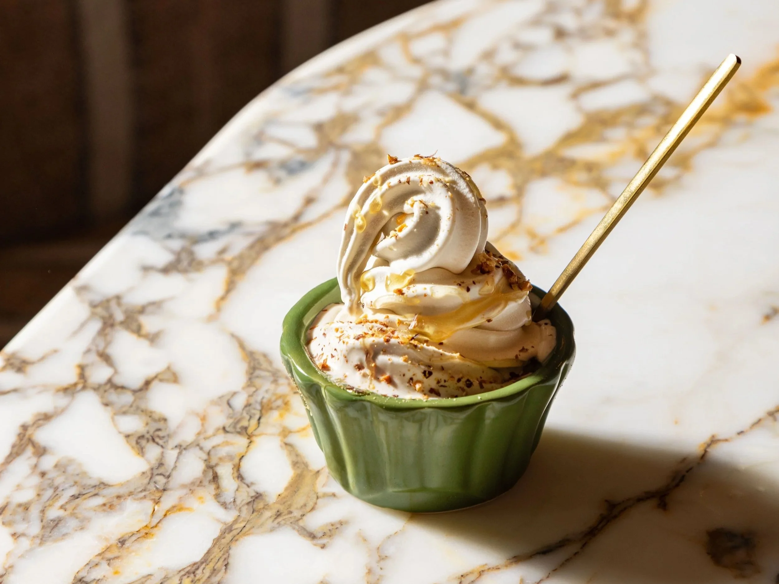 A small green ceramic cup filled with soft serve ice cream topped with a swirl of vanilla ice cream and sprinkles, placed on a white marble table with gold and gray veining.