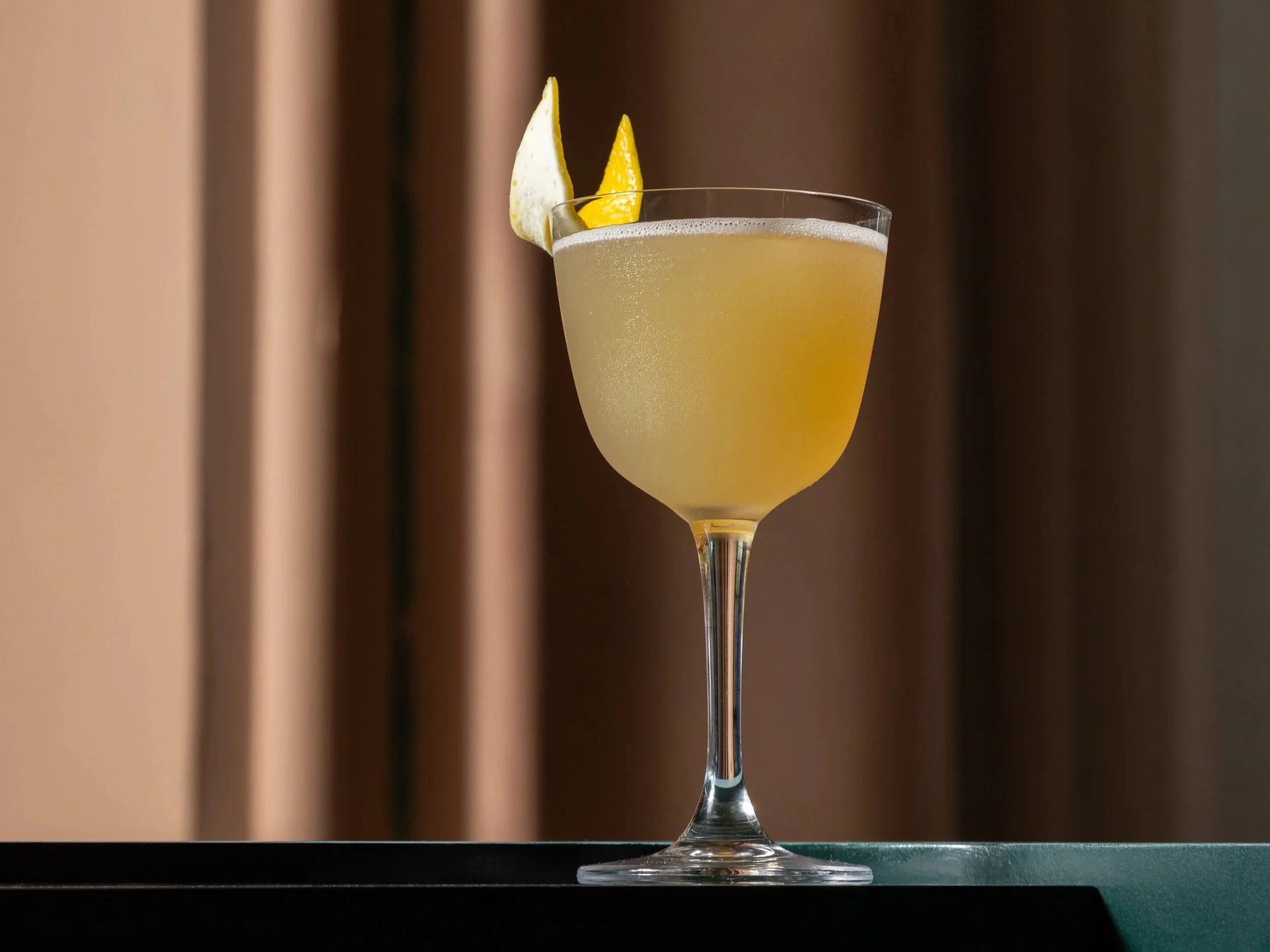 A cocktail in a stemmed glass garnished with lemon and lime wedges against a blurred background