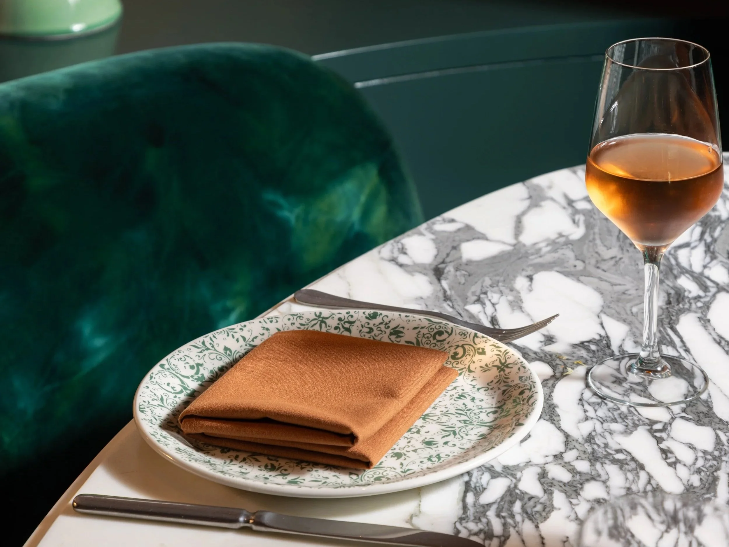 A marble table set with a patterned plate, a brown folded napkin, a knife, a wine glass with rosé wine, and a green velvet chair. There is a green vase and a lamp in the background.
