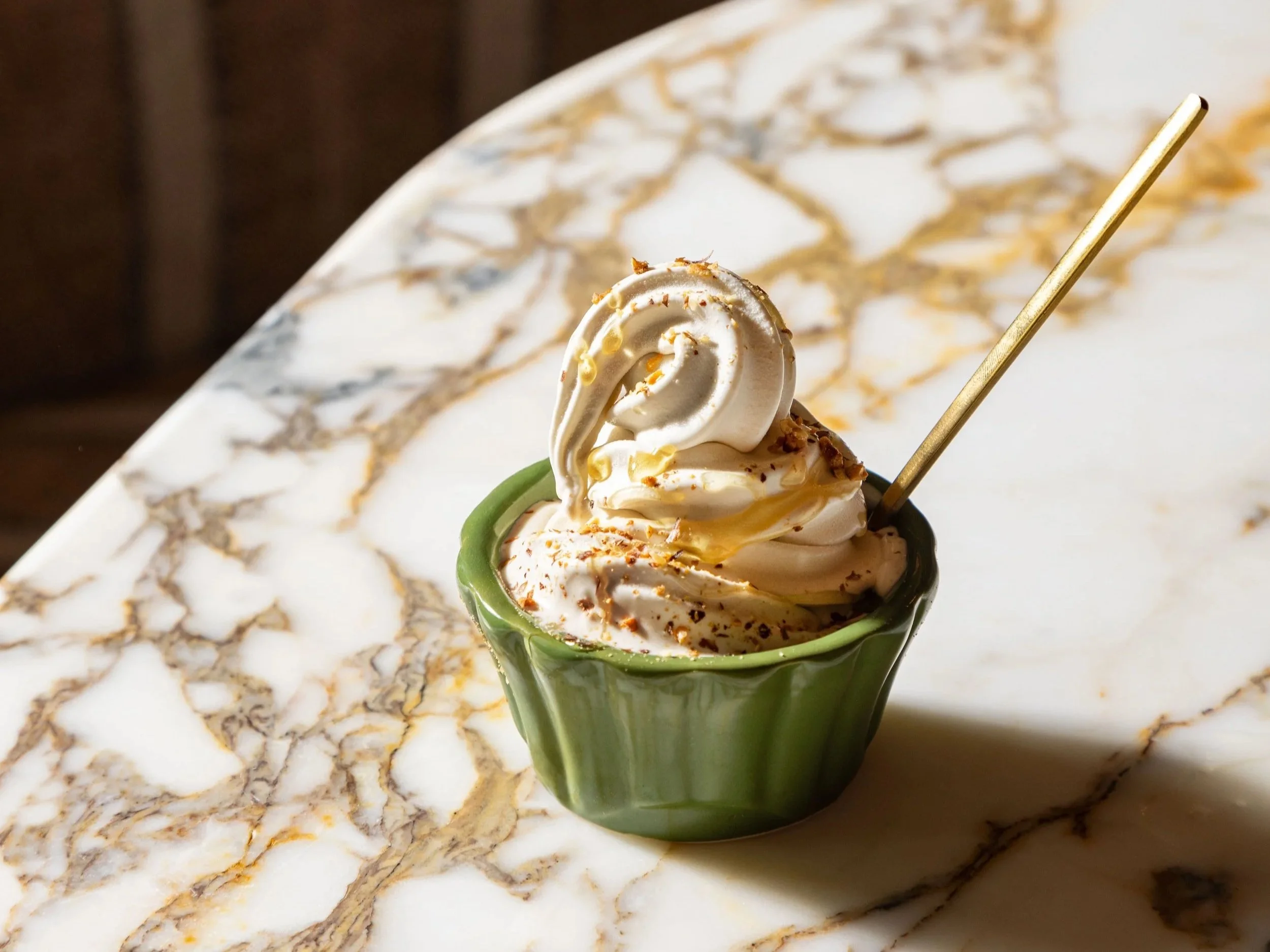 A small green ceramic cup filled with soft serve ice cream topped with a swirl of vanilla ice cream and sprinkles, placed on a white marble table with gold and gray veining.