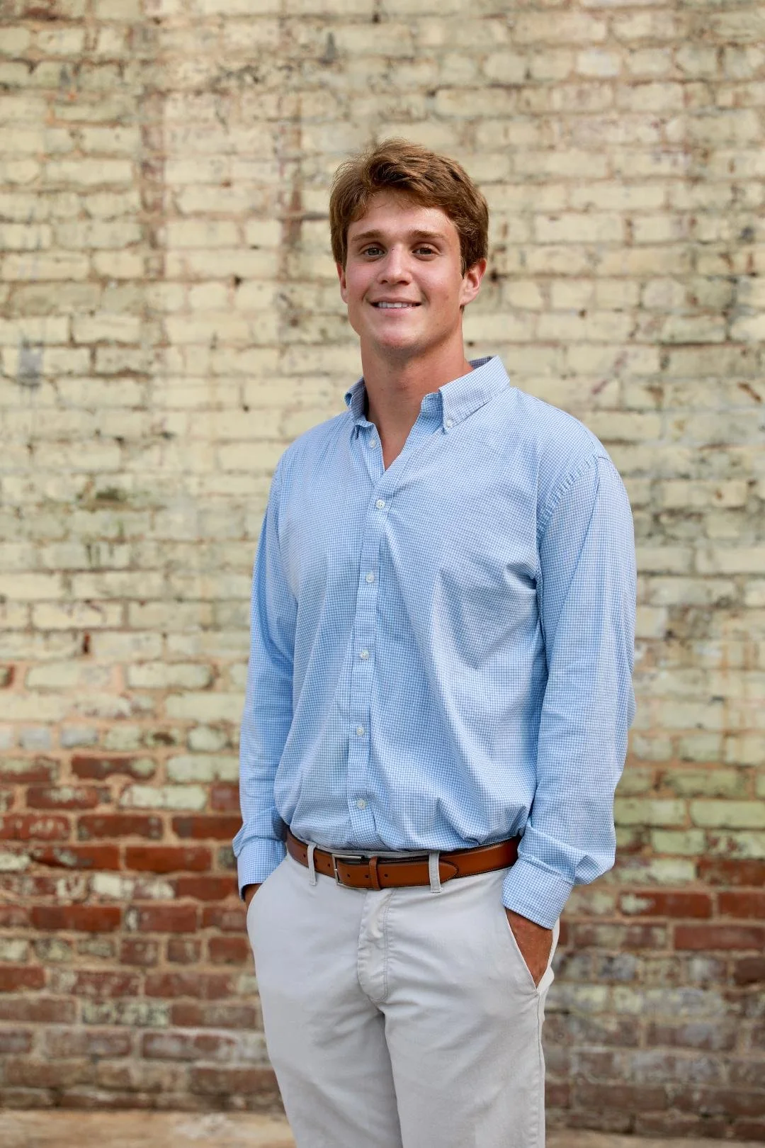 A young man with light skin and brown hair standing in front of a brick wall, wearing a light blue button-down shirt and beige pants, smiling at the camera with his hands in his pockets.
