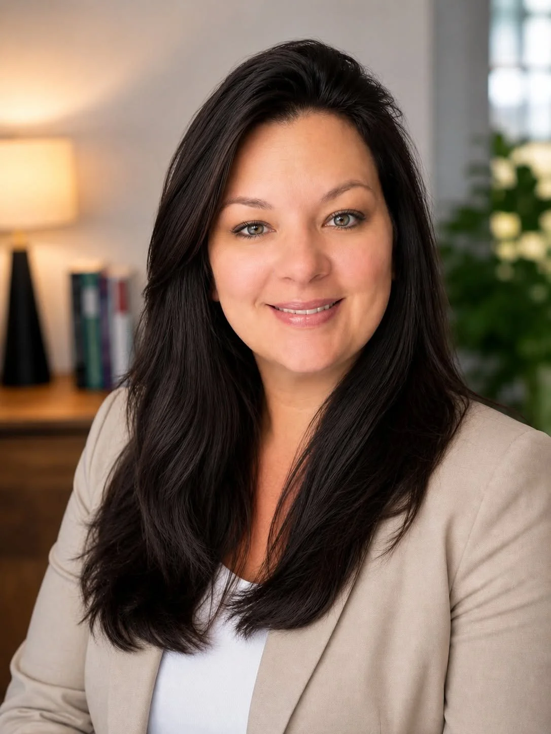 A woman with long dark hair and light skin smiling, wearing a beige blazer and white top, in an office or home setting with a lamp, books, and a green plant in the background.