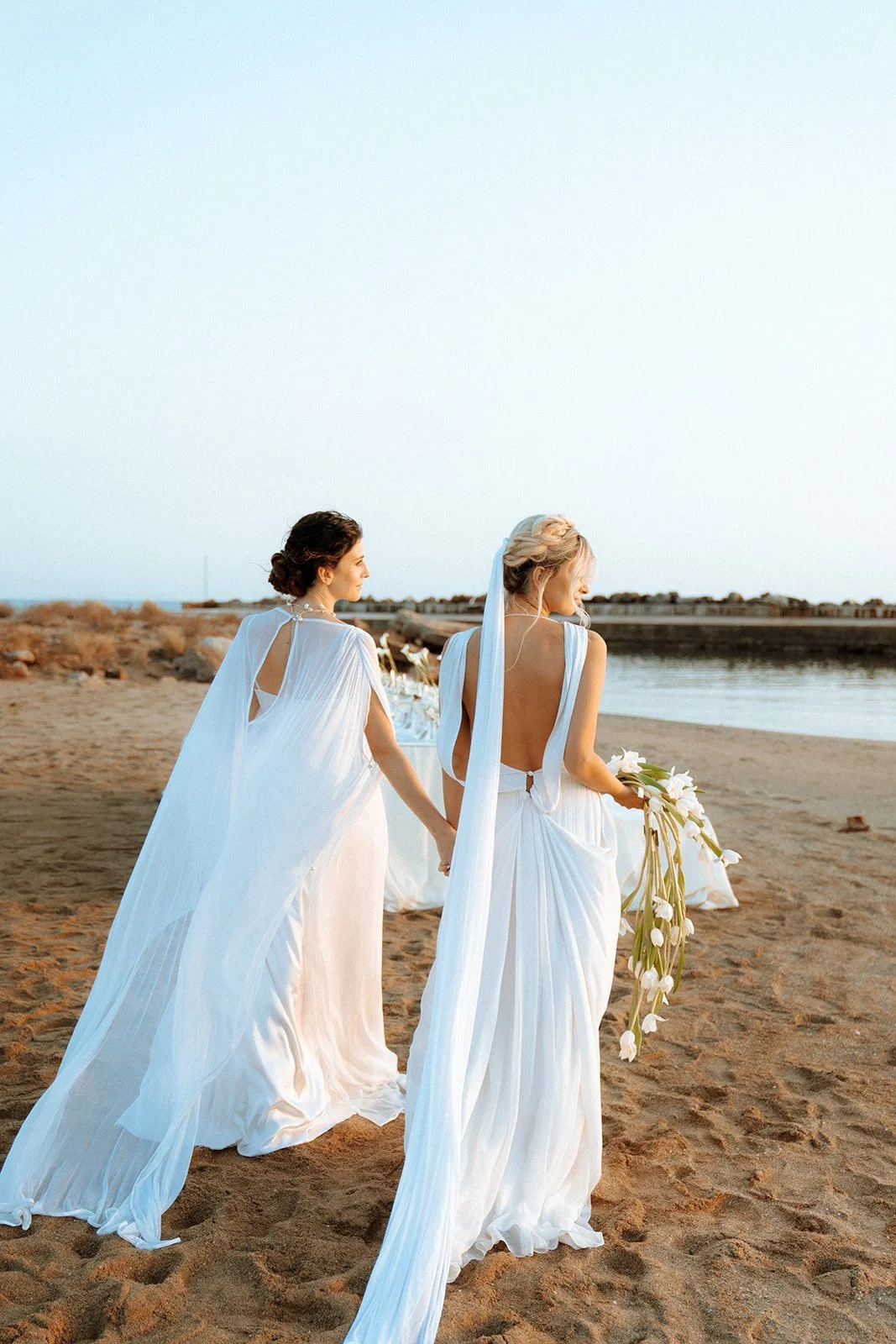 2 brides walking on the beach holding hands with cape and veil on