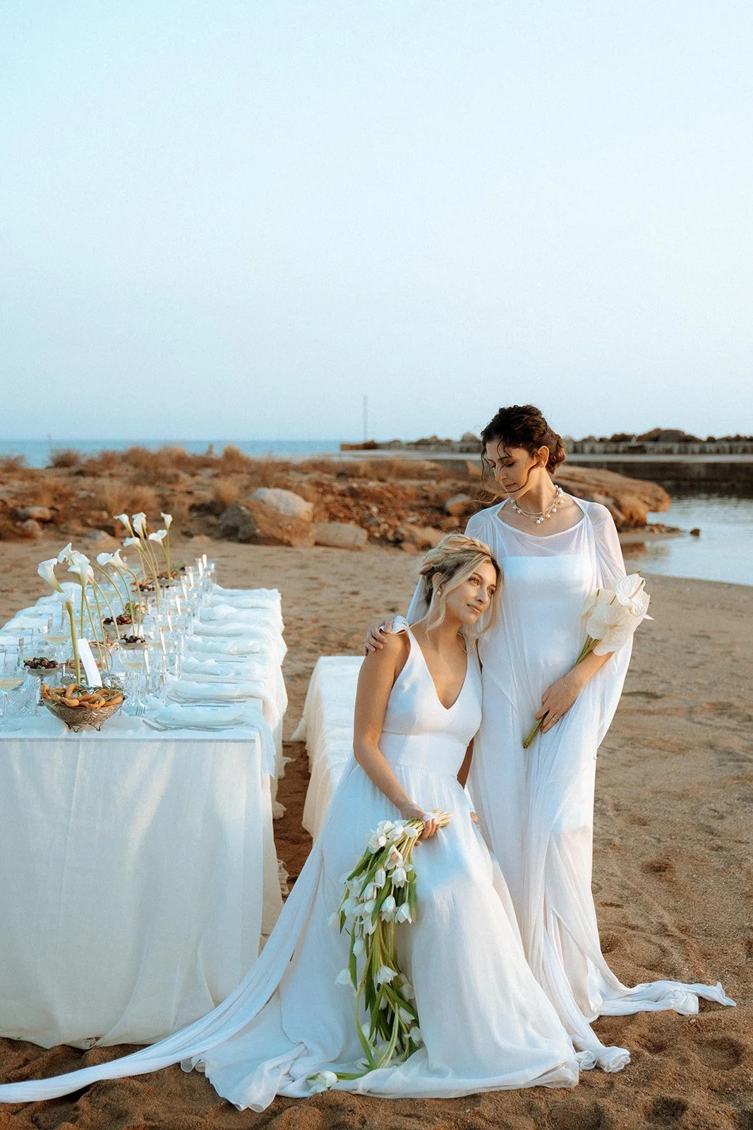one bride standing and one bride sitting, holding bridal bouquets next to a table decor 