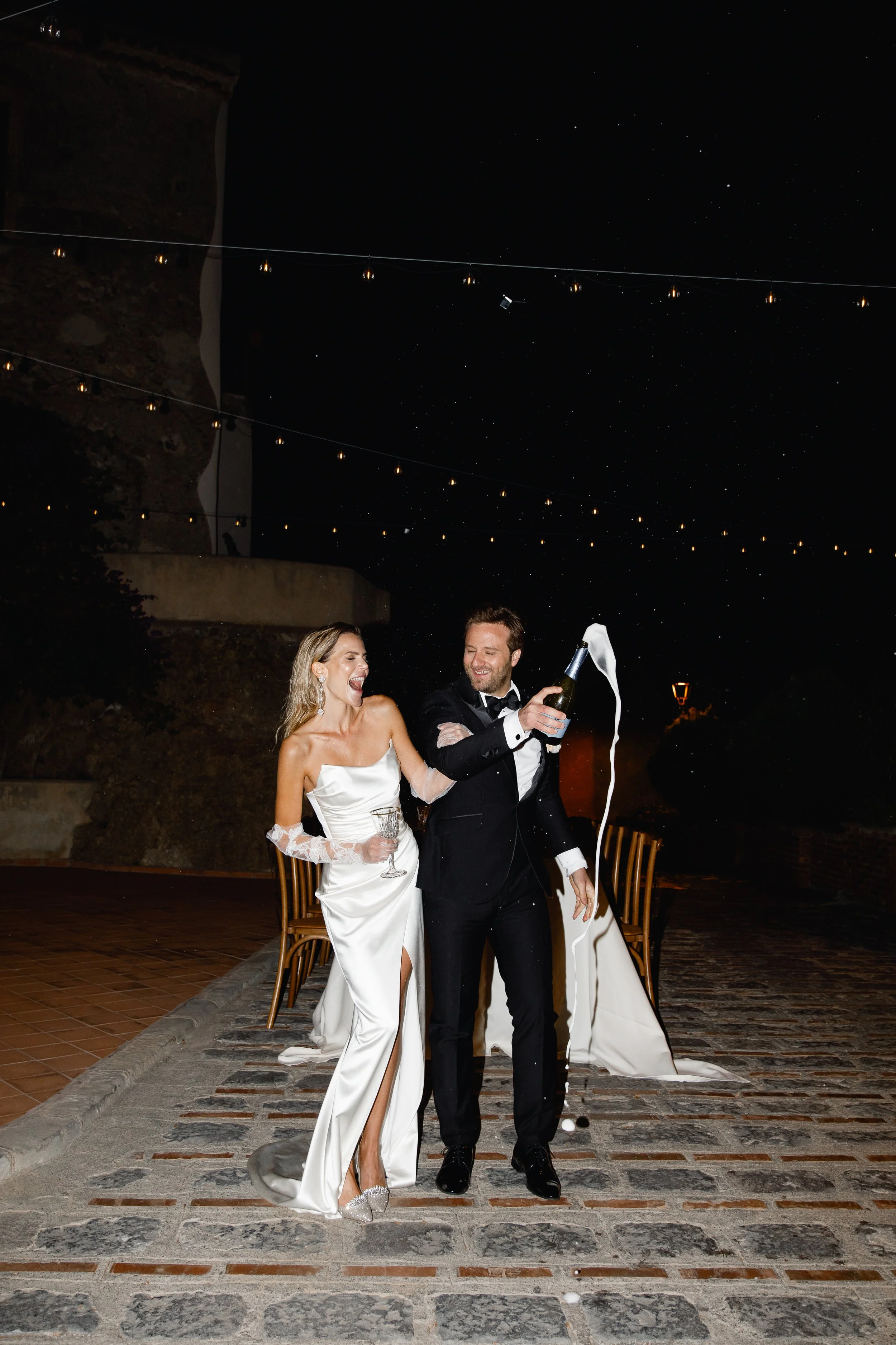 A newlywed couple celebrating at night, with the groom opening a bottle of champagne, causing it to overflow, while the bride laughs and holds a glass of champagne. They are dressed in formal wedding attire, standing outside on a paved area with string lights overhead under a starry sky.