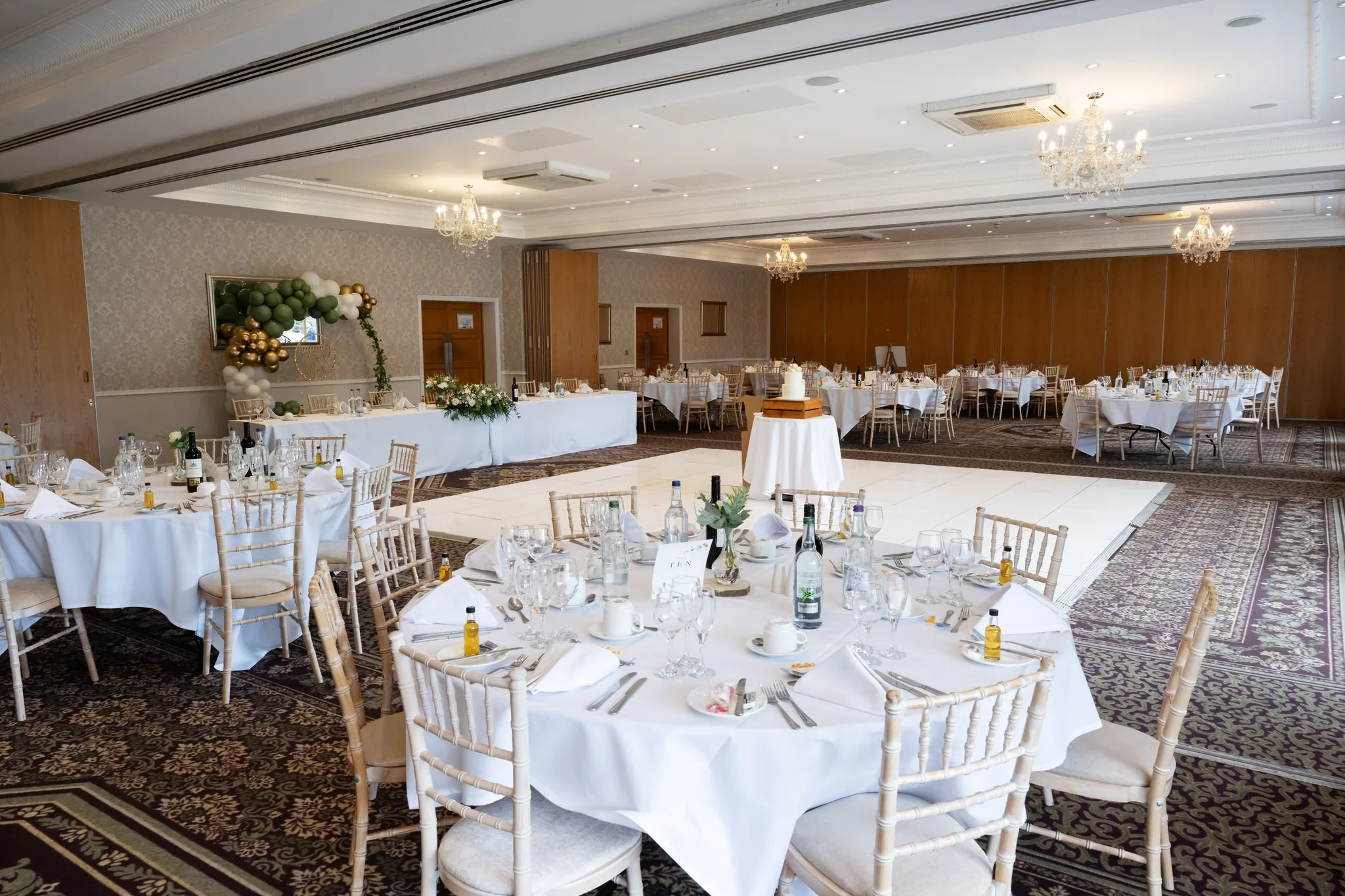 Elegant banquet hall decorated for a wedding reception with round tables, white tablecloths, chairs, floral centerpieces, candles, and a cake on a stand in the center.