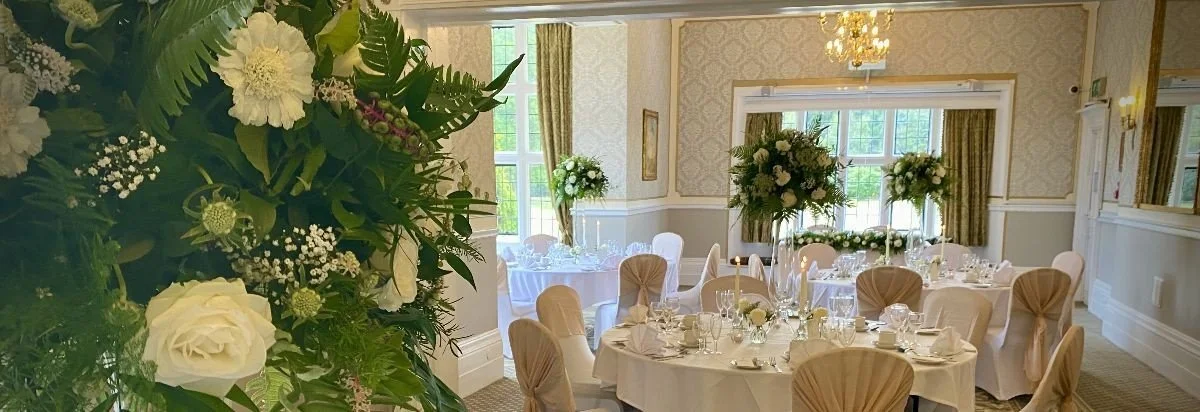 Elegant banquet hall decorated for a wedding reception with tall floral arrangements, round tables with white tablecloths, and beige chair covers in a well-lit room with large windows and gold curtains.