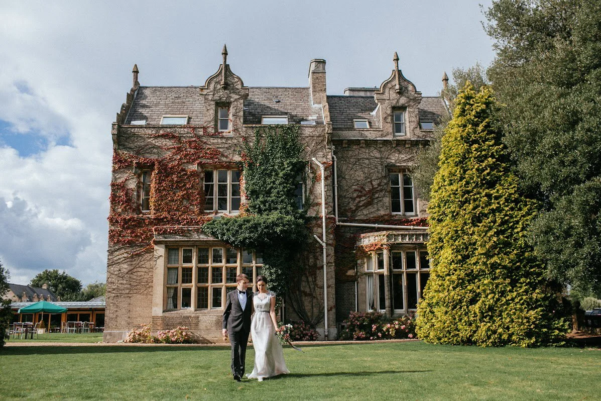 Wedding couple walking on grass in front of a large historic brick house covered in vines with lush trees around.