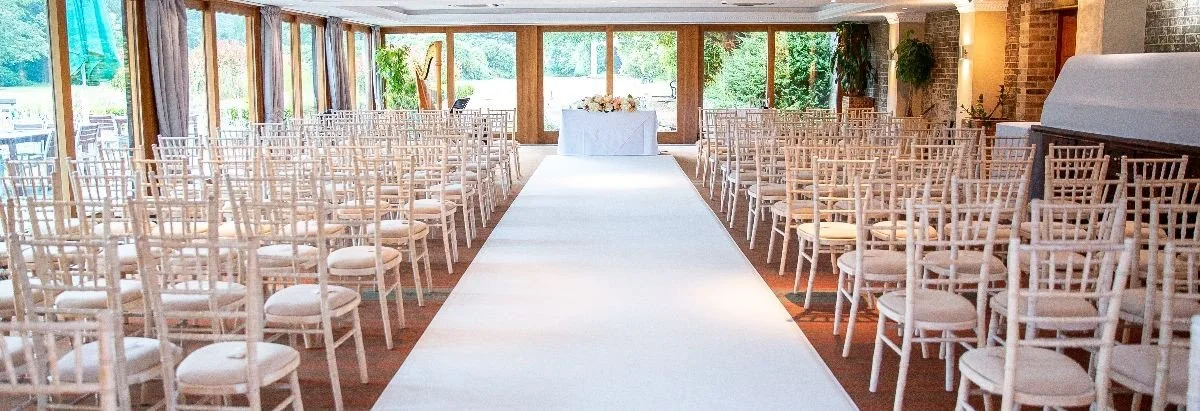 Empty wedding ceremony aisle with white chairs on either side and a flower-decorated table at the front of the room, large windows letting in natural light, and greenery outside.