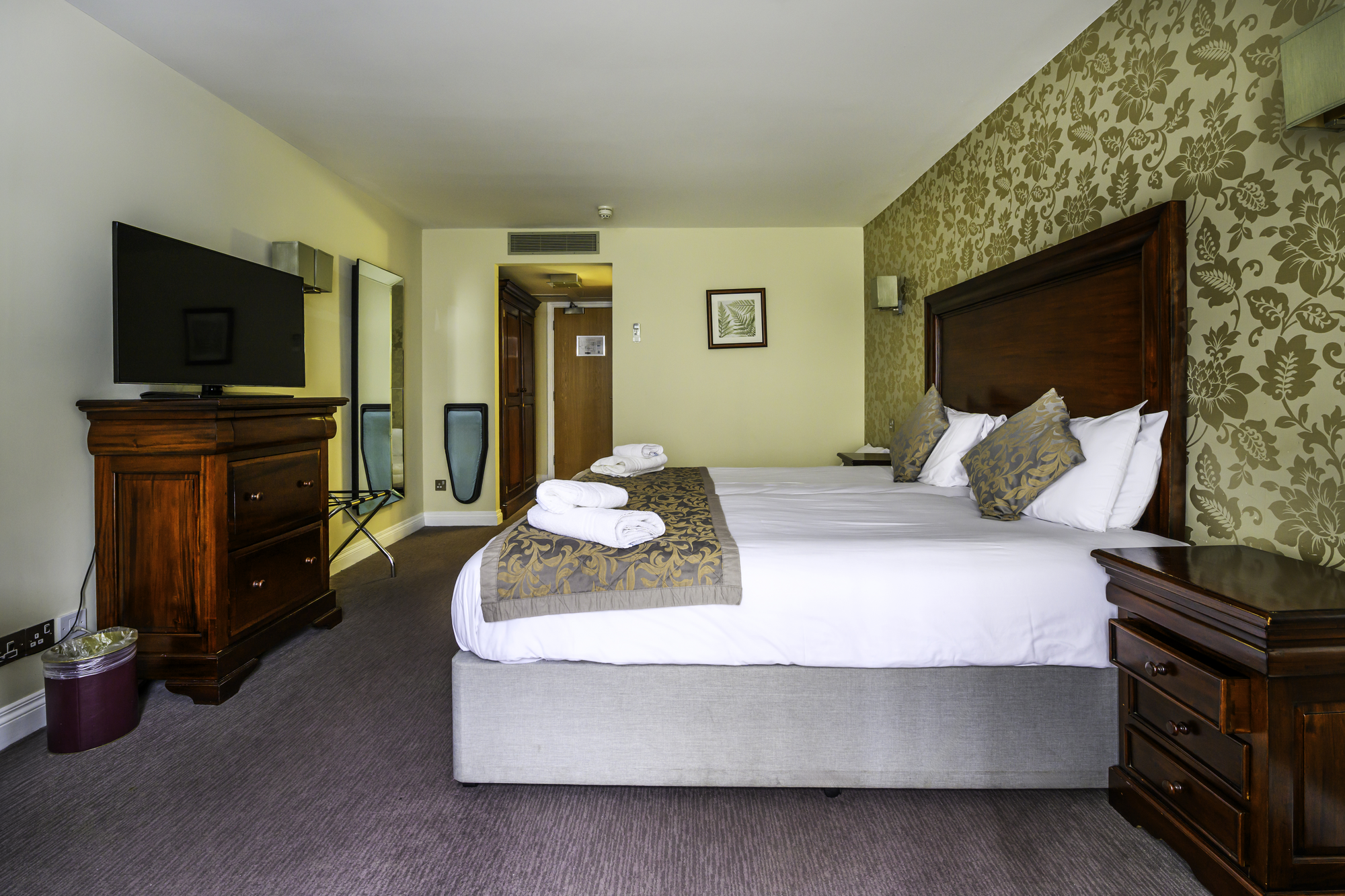 A hotel room with a large bed, wooden headboard, and matching nightstands, with decorative pillows and folded towels on the bed, a flat-screen TV on a wooden dresser, and a mirror and artwork on the walls.