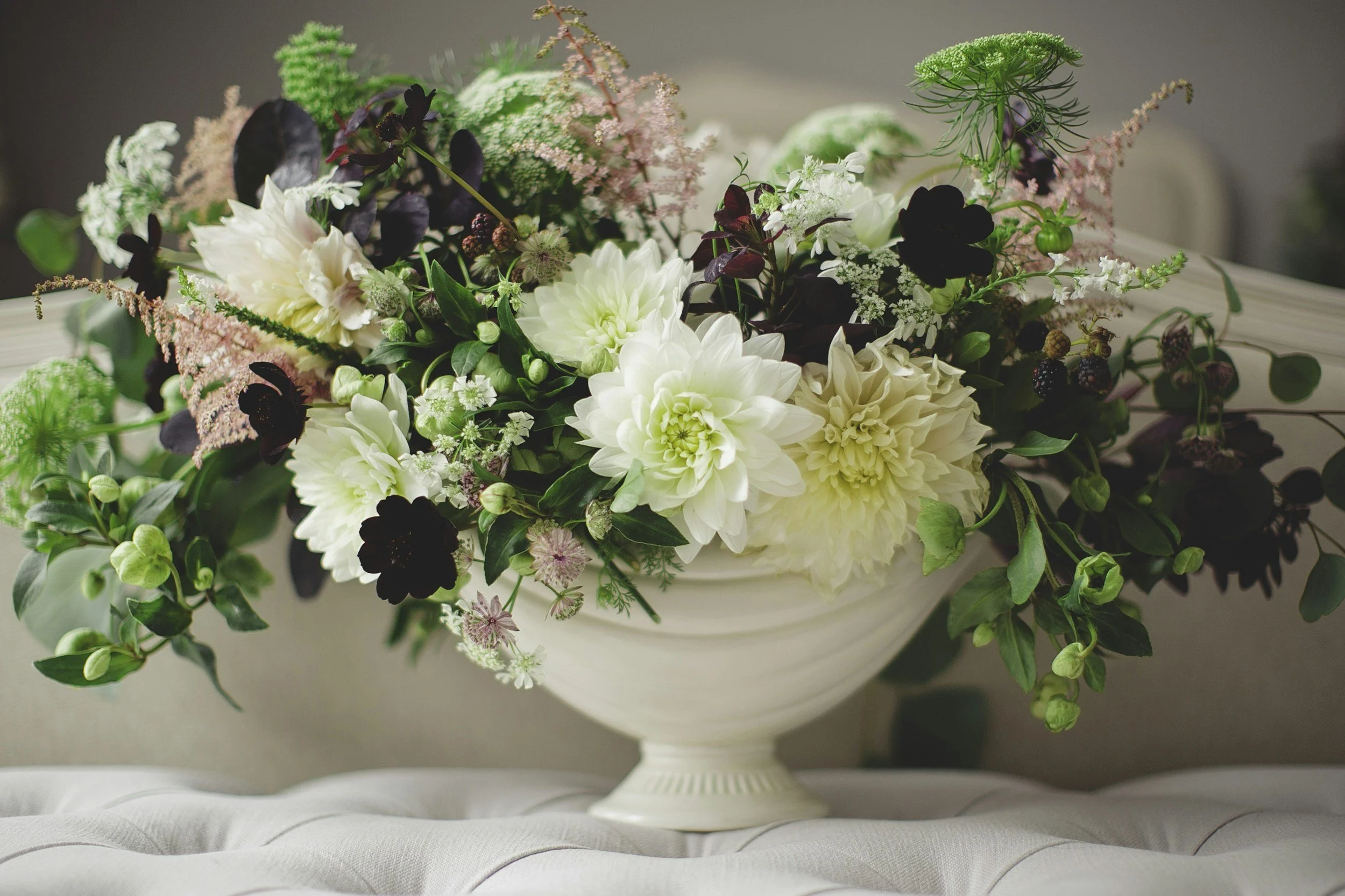 A decorative floral arrangement in a white vase featuring white dahlias, dark purple and black flowers, pink astilbe, and various green leaves and berries, placed on a cushioned surface.