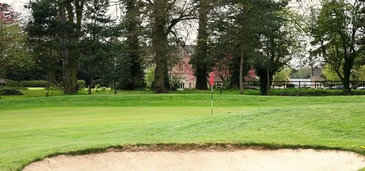 A golf course with a sand trap in foreground, green fairway, trees, and a flag on the green, with houses in background.