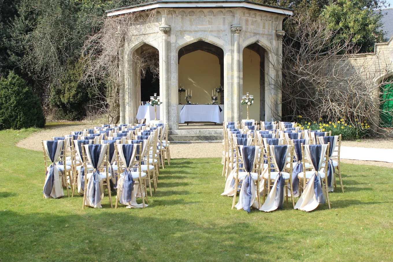 Wedding ceremony setup outdoors with rows of decorated chairs facing a small stage with a table, candles, and floral arrangements in front of an ornate stone structure surrounded by trees and shrubs.