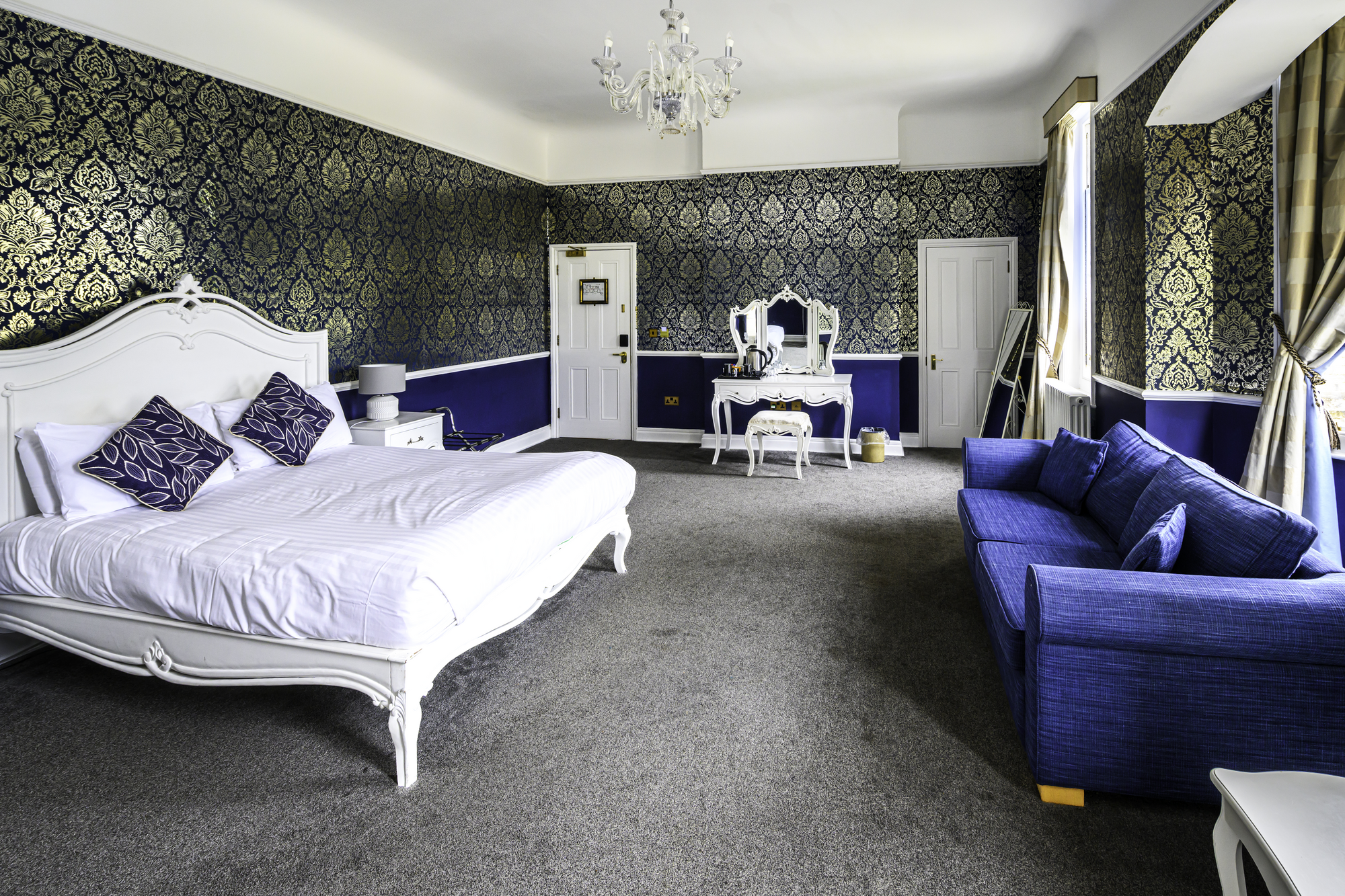 A luxurious hotel hotel room with a white bed, navy blue accents, patterned wallpaper, and a seating area with a navy blue sofa.